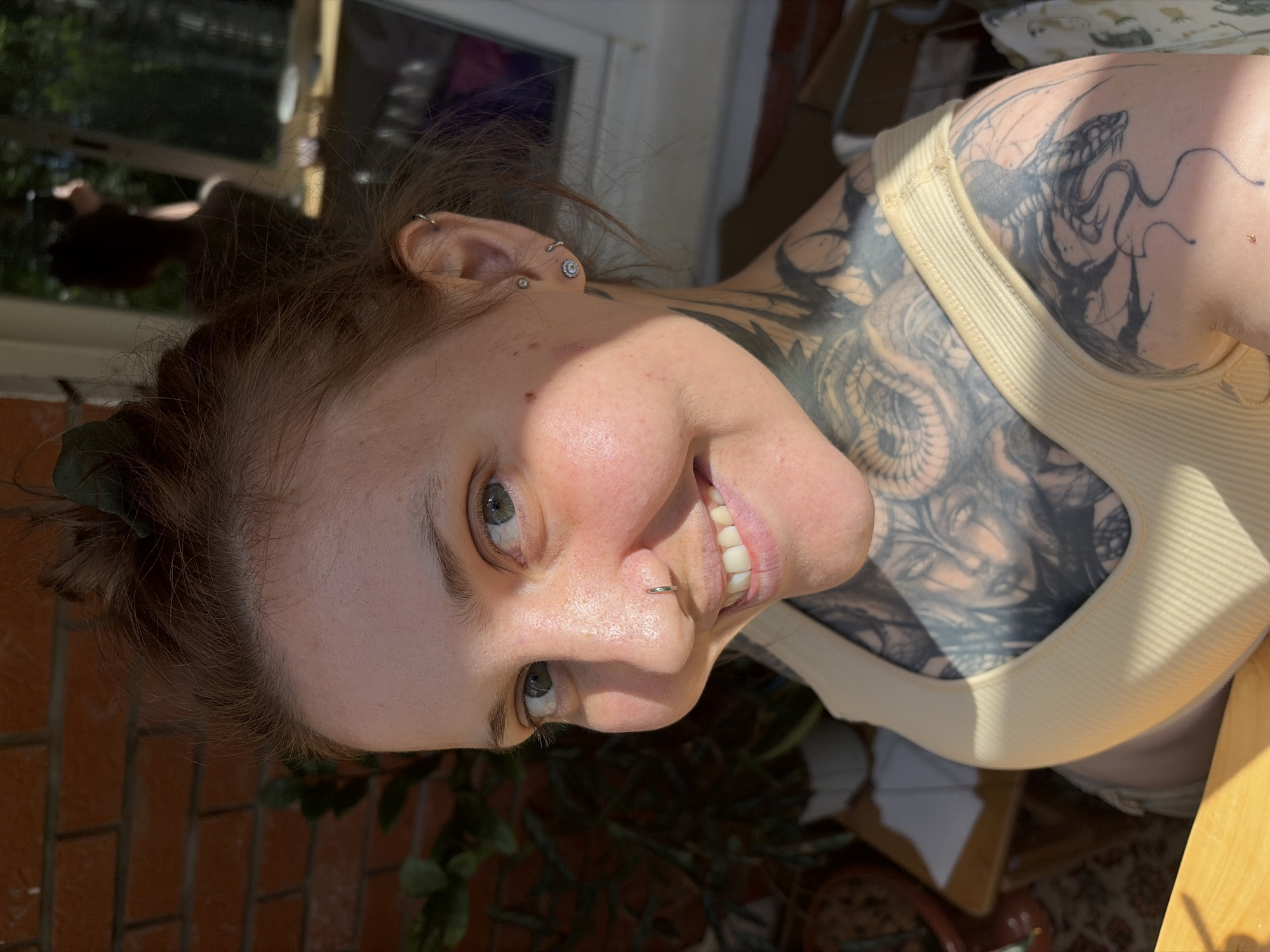 A close-up, sideways portrait shows a woman with reddish-brown hair, fair skin, and green eyes. She is smiling, revealing her teeth, and has a nose ring. Several piercings are visible on her ear, including a stud and a hoop. The woman has extensive tattoos on her neck and shoulder, featuring intricate designs, including what appears to be a dragon. She is wearing a ribbed, cream-colored tank top. The lighting suggests it is daytime, with sunlight casting shadows on her face. The background includes hints of greenery, brickwork, and furniture, suggesting an indoor or patio setting. The overall mood is relaxed and happy. No visible text is present in the image.