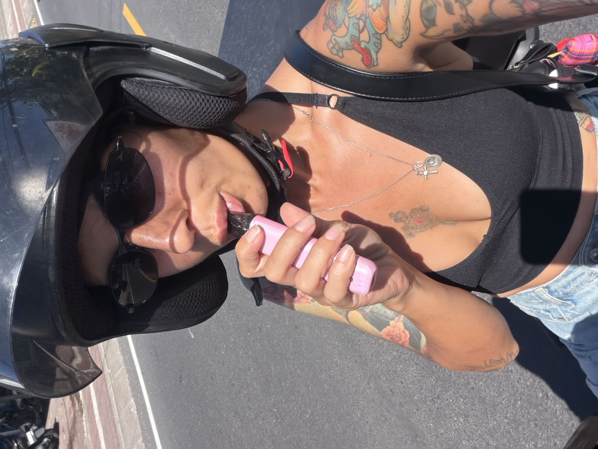 In Kuta Selatan, Indonesia, a person wearing a black helmet and sunglasses is on a motorcycle, holding a pink vape device to their mouth. They have tattoos on their arms and chest and are wearing a black tank top and denim shorts. The road behind them is paved with a yellow line marking.