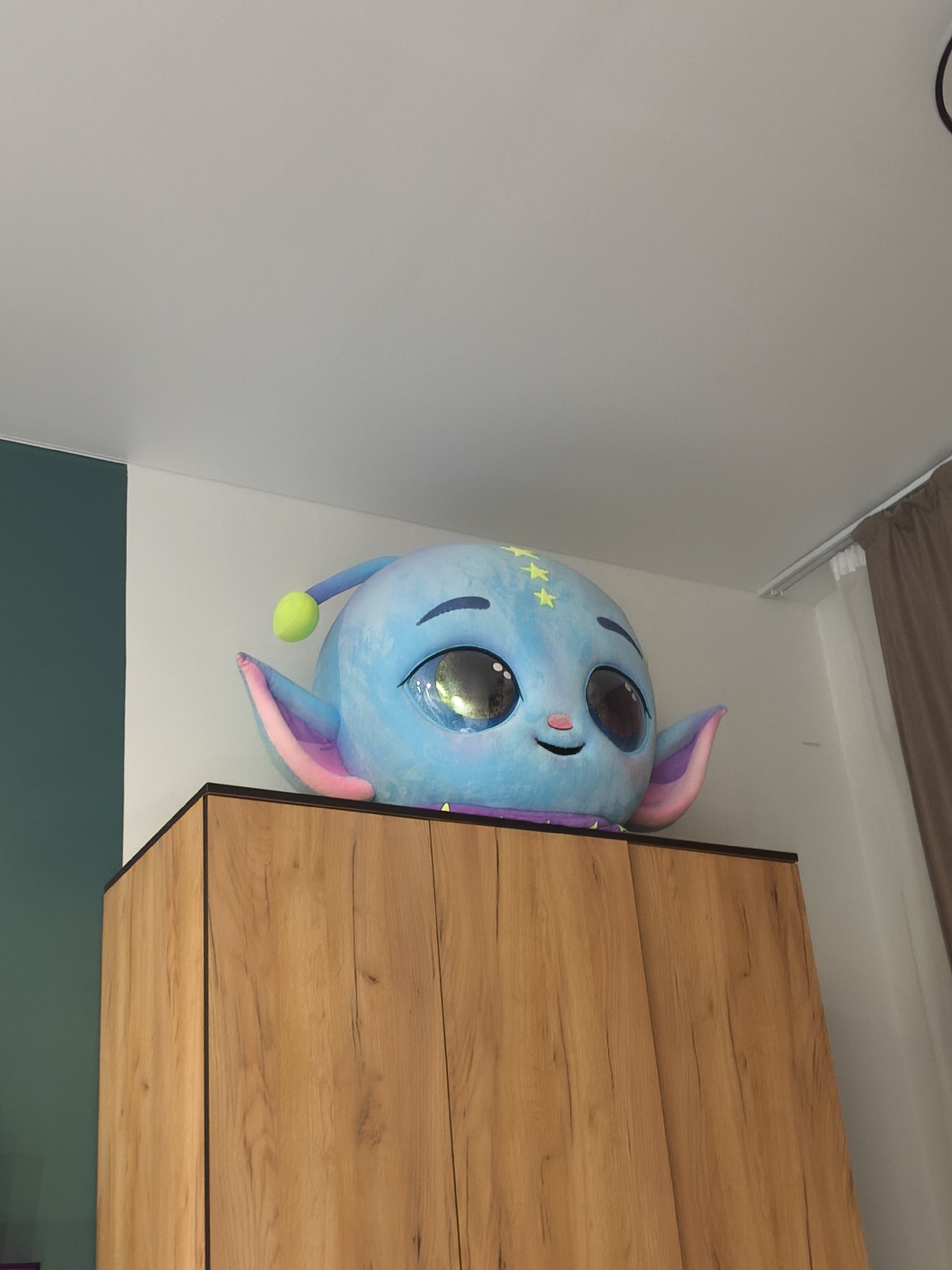 A blue plush alien-like toy with large eyes, a small smile, and one antenna sits atop a wooden cabinet. The cabinet has a faux wood grain pattern and dark trim. The background consists of a white wall, a section of green wall, and a brown curtain. The ceiling is white. The location is in a room within an unspecified building, and there are no indications of the city of Moscow, Russia.