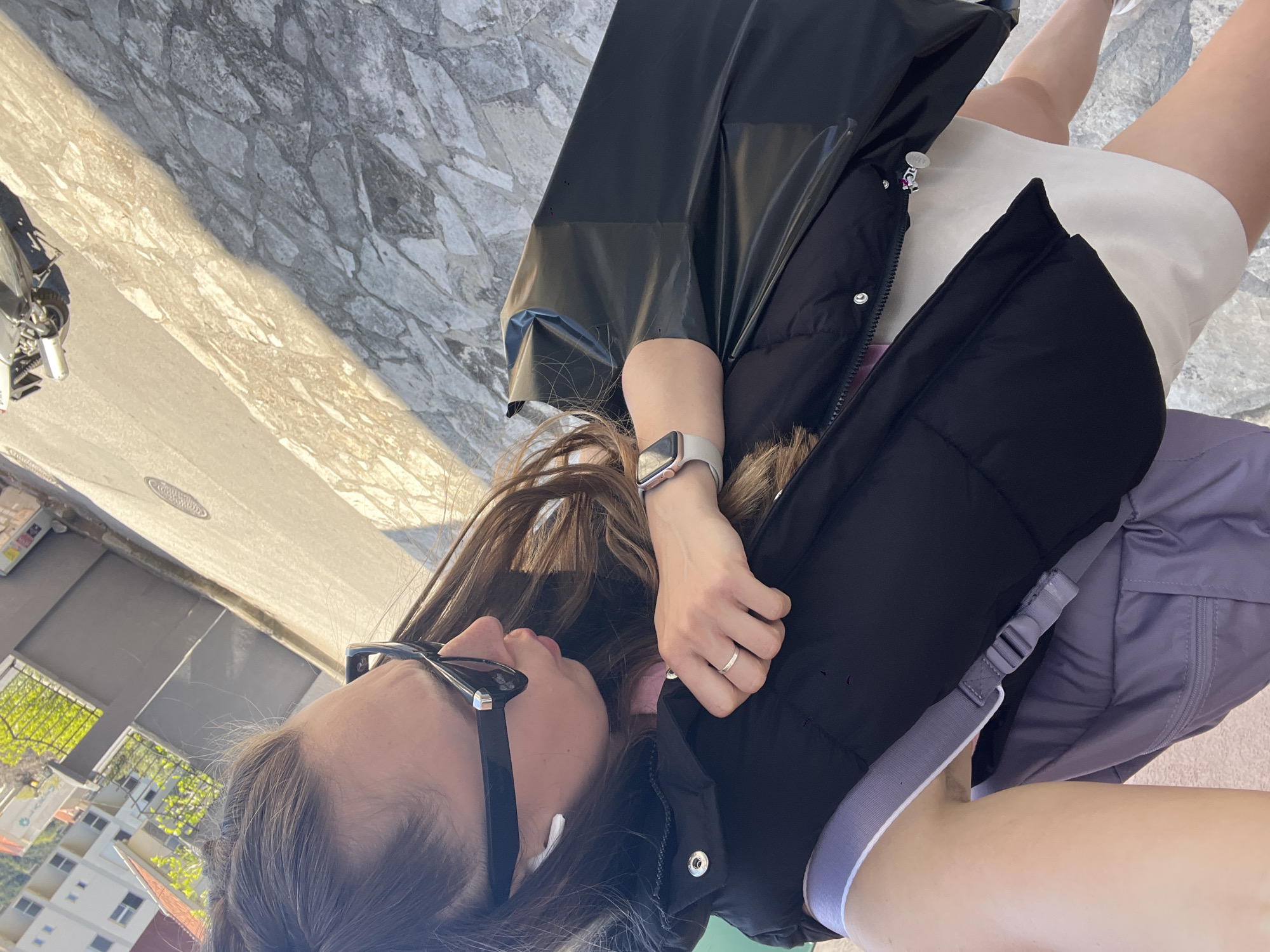 A woman is depicted in a tilted, close-up shot. She wears sunglasses, an earpiece, a black puffer vest, a light-colored dress, and a watch. A ring adorns one of her fingers. Part of a shiny black garment or bag is also visible. A greyish-purple backpack is partially visible. The background reveals a street scene with buildings and greenery, potentially indicating an urban area in Bečići, Montenegro. The street itself has a mix of paved and cobblestone surfaces.