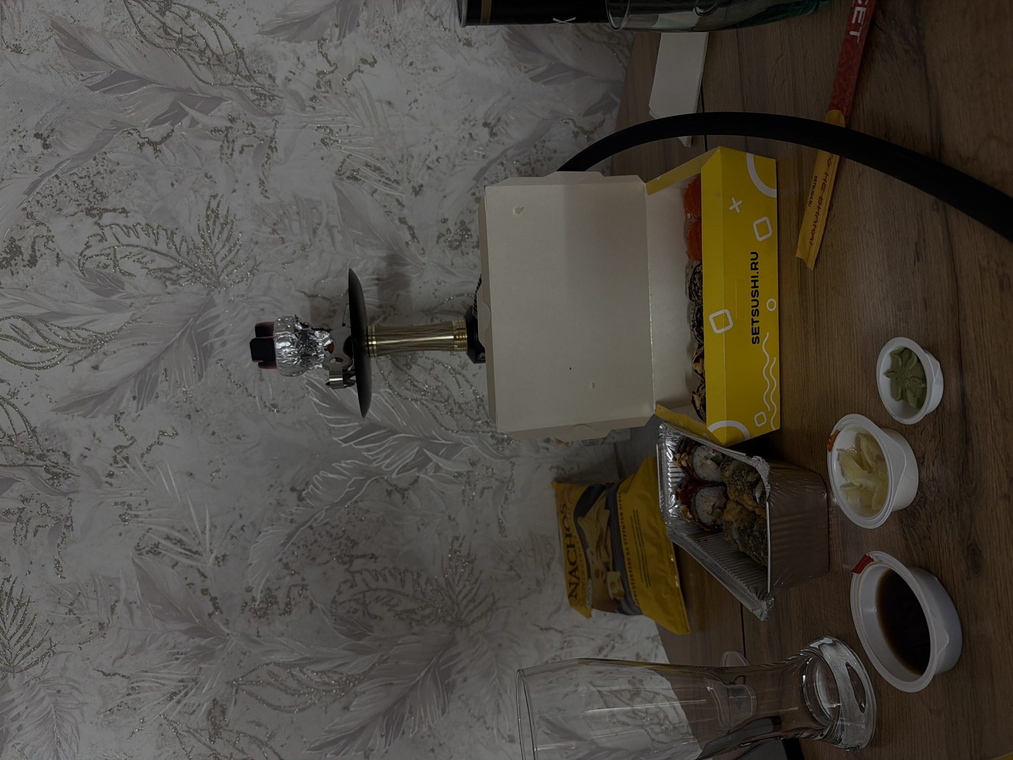 The image shows a tabletop scene, likely indoors, with a variety of items. Sushi in a box and a foil container are present, along with condiments (wasabi, ginger, soy sauce). A bag of Nachos and a hookah are also visible. A glass stands on the table. Based on the "SETSUSHI.RU" branding and the presence of the hookah, the scene appears to be a casual meal setting. There is no information in the image that indicates this scene is located in Reshetnikovo, Russia. The wall has a decorative wallpaper pattern.
