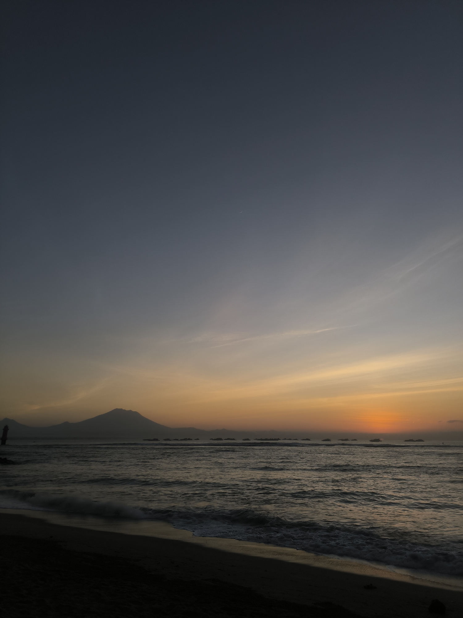 This is a vertical, eye-level photograph of a sunset scene at a beach in Denpasar, Indonesia. The sky is a gradient of deep blue at the top, transitioning to pale yellow and orange near the horizon. A faint crescent moon is visible in the upper left quadrant of the sky. Wispy clouds are scattered across the sky, catching the warm colors of the sunset.
In the mid-ground, the dark silhouette of a mountain range is visible across the water. The ocean stretches from the foreground to the horizon, with gentle waves lapping onto the shore. Several small boats are dotted across the water in the distance, appearing as dark shapes against the brighter sky. A lighthouse-like structure is faintly visible on the left side of the image.
The foreground consists of a dark sand beach. The overall mood is serene and peaceful, characteristic of a sunset at the coast. There are no people or animals visible in the scene. The image captures the natural beauty of the coastal landscape during the twilight hours.