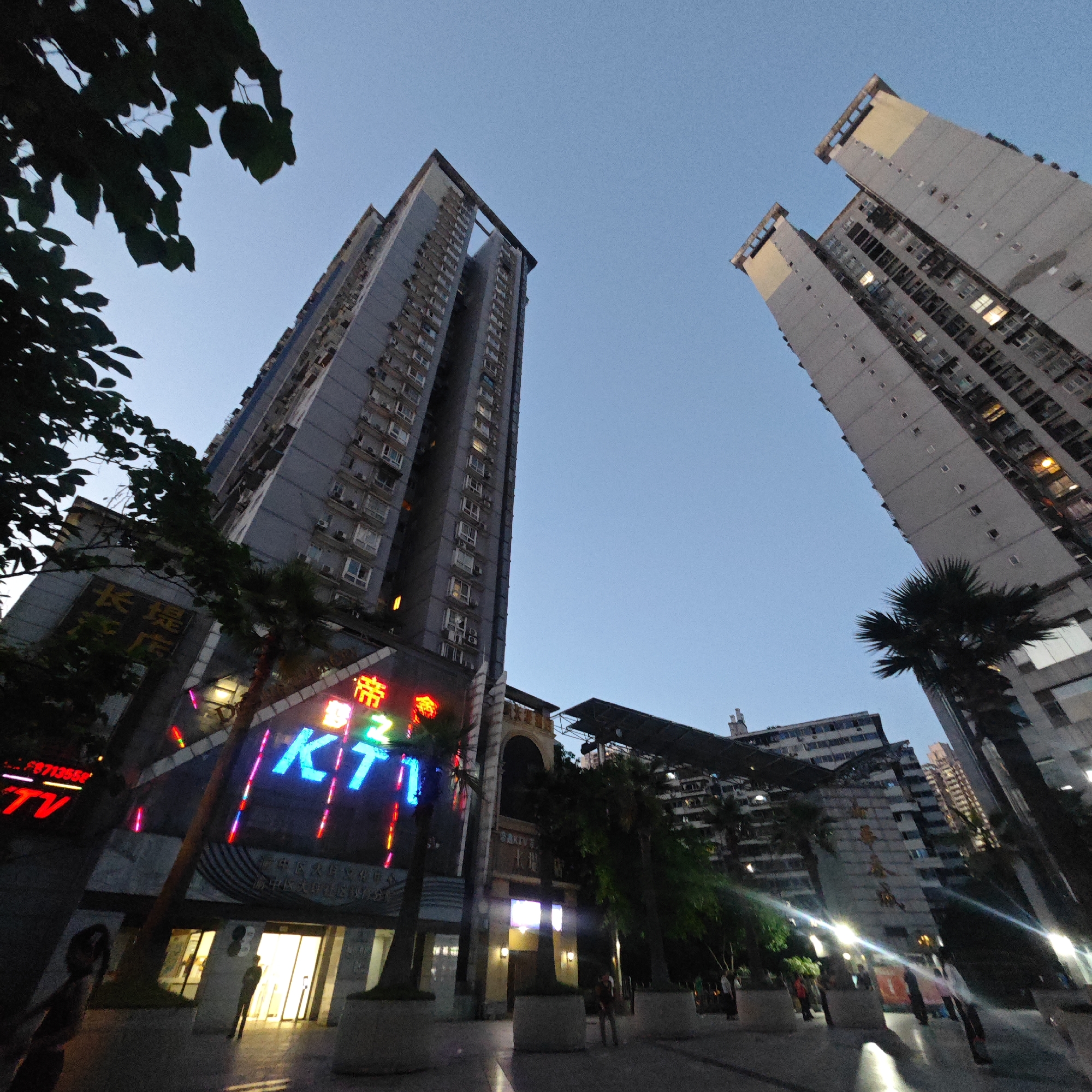 The image is a low-angle shot of a street scene in the Yuzhong District of China, with a focus on tall buildings and street-level commercial spaces. The sky is a clear blue transitioning into twilight.
To the left is a multi-story building with "长堤大飯店" inscribed in gold above its entrance, flanked by neon signs reading "TV" in red and "KT" in blue. The building's façade is modern, with a geometric design and palm trees planted in front. A few people are walking in and out of the brightly lit entrance.
Further back and to the right stands a taller, residential building with multiple windows and a light grey exterior. Palm trees are also present in front of this building. Between the two buildings, other structures and buildings can be seen, including one that appears to have an open-air roof structure.
The ground is paved with light-colored stone tiles, reflecting light from various sources. There are people walking on the sidewalk, adding a sense of activity to the scene.
