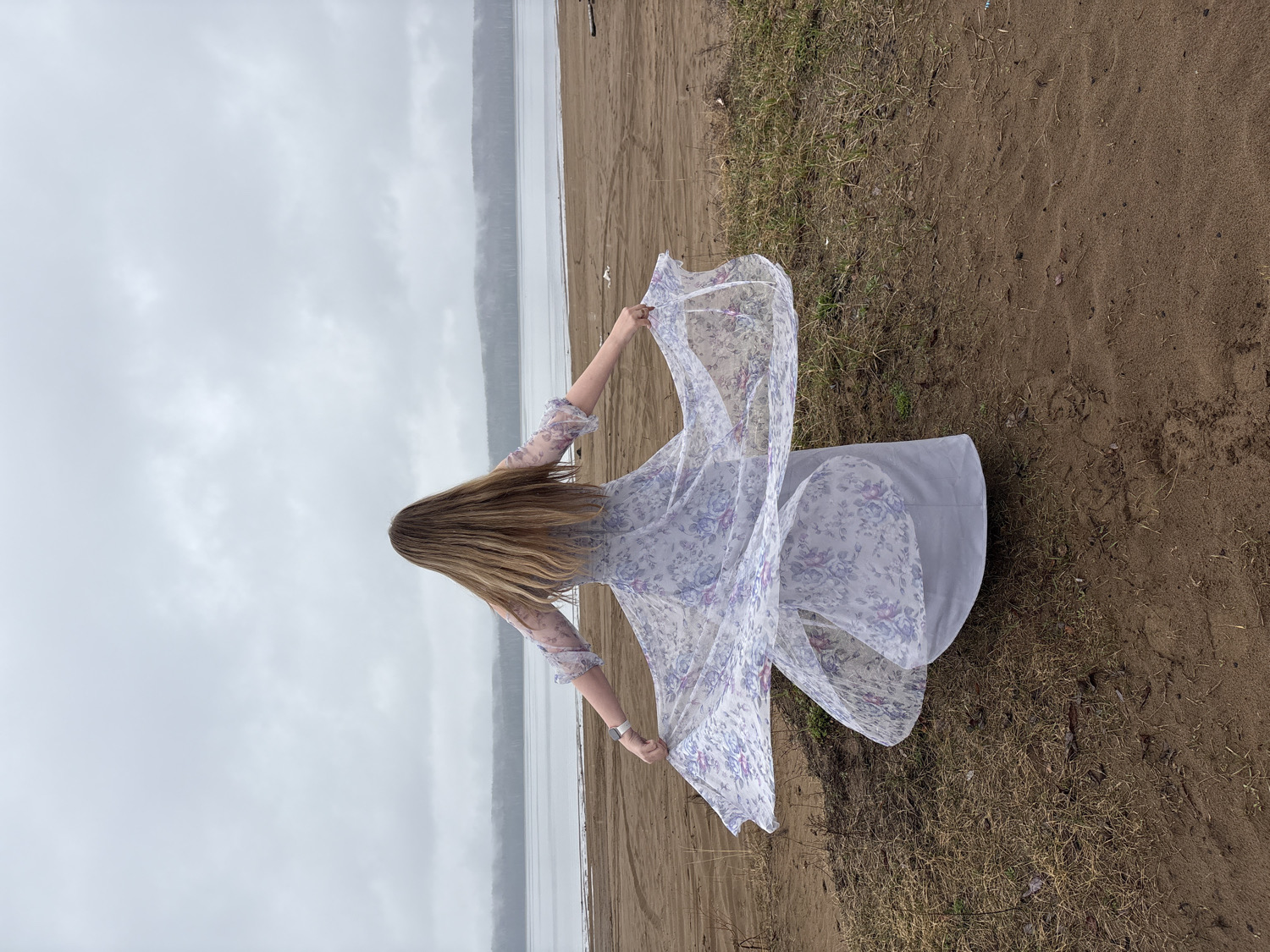 The image displays an outdoor scene of a person on a sandy or muddy shoreline next to a wide body of water, possibly a lake or a large river. The sky is uniformly overcast and gray, indicating a cloudy day.
The primary subject is a person, likely a woman, viewed from behind, positioned towards the right side of the frame. She has long, straight, light brown hair flowing down her back. She is wearing a long, flowing dress that is predominantly white or light gray, patterned with delicate purple and possibly blue floral designs. Her arms are outstretched to her sides, holding the sheer, outer fabric of her dress, which billows dramatically around her, suggesting movement such as twirling or dancing. A silver watch or bracelet is visible on her left wrist.
The foreground consists of dark brown sand or mud, interspersed with patches of dry grass. In the mid-ground, the water body extends horizontally, meeting the distant horizon where faint, indistinct landmasses or hills are visible through the hazy, overcast conditions. The overall impression is one of freedom, grace, and contemplation amidst a natural, somewhat subdued landscape. There is no visible text in the image.