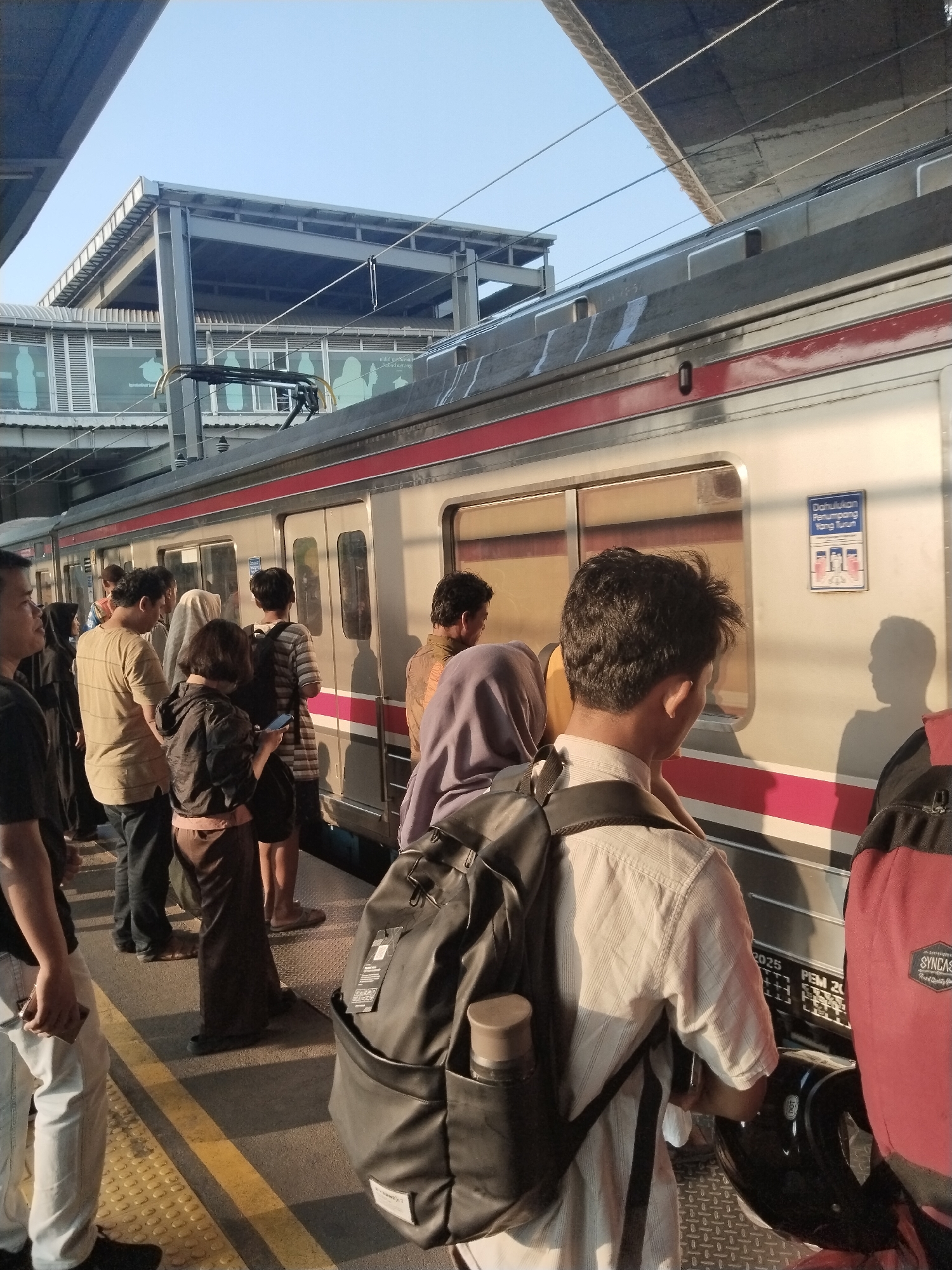 A group of people are standing on a train platform in Central Jakarta, Indonesia, waiting for a train. The train is silver with red and pink stripes along the side. There are overhead structures and a clear sky above. The people are dressed in casual clothing, and some are carrying bags and backpacks. One person in the foreground is looking at their phone. There is a blue and white sign on the side of the train with text and images of people. The platform has yellow lines indicating the edge.