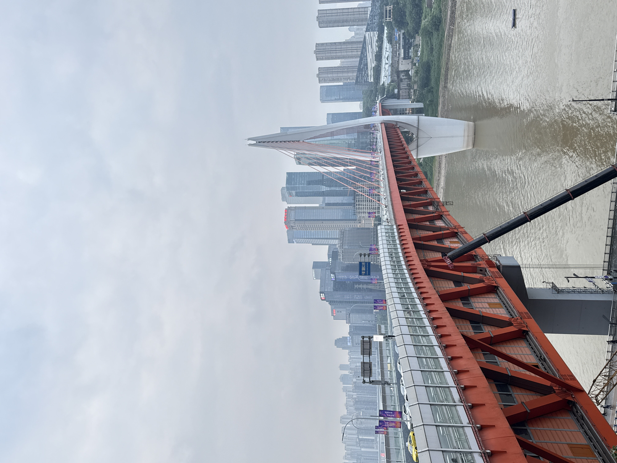 The image shows a view of the Hongya Dong Bridge in Chongqing, Yuzhong District, China. The bridge, which is painted a vibrant orange, spans a wide, muddy river. Its unique design features a large white pylon from which numerous cables extend to support the bridge deck. In the background, a dense cityscape of modern skyscrapers rises against a cloudy sky. Cars and traffic lights are visible on the bridge's upper deck, indicating it is a functional transportation route. The river below appears calm, with a small boat visible in the distance. The overall impression is one of urban development and infrastructure in a large Chinese city.