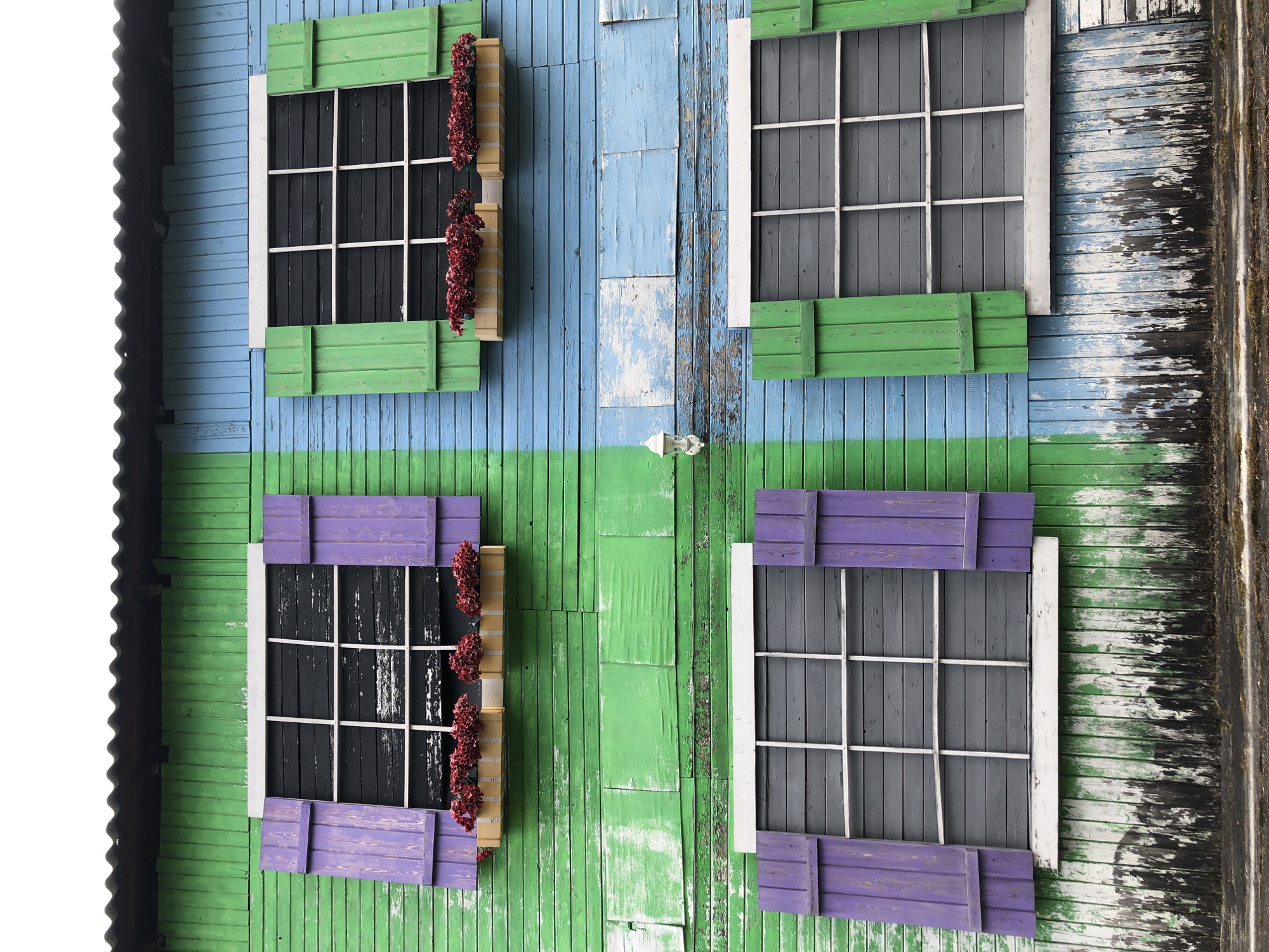The image shows a close-up view of a building facade in Krasnoyarsk, Russia. The wall is constructed of vertical wooden planks and painted with alternating sections of blue and green paint. The paint is aged and peeling in places. There are four rectangular windows arranged in two rows of two. The windows have white frames and are covered with wooden shutters, painted green on the top row and purple on the bottom row. Two of the windows have flower boxes attached to them, overflowing with red flowers. The windows themselves are black. To the left is a white corrugated roof.