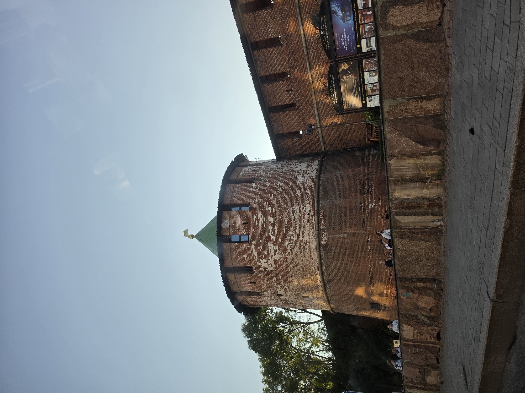 The image depicts a section of the Nizhny Novgorod Kremlin, specifically focusing on the Dmitrovskaya Tower and a portion of the adjoining wall. The tower, constructed of red brick, has a conical green roof topped with a small golden finial. The wall is also made of red brick, with crenellations along the top. In front of the wall, there is a stone embankment with several people standing and sitting along it, facing towards the Kremlin. The embankment is made of large, rough-hewn stones in shades of brown and grey. A paved walkway runs in front of the embankment, and a street is visible beyond that. Two large posters are mounted on the wall behind the embankment, advertising events for the "Filarmoniya Rostopcha" (Rostopcha Philharmonic). The sky is clear and pale blue. Trees are visible to the left of the tower, and the scene is illuminated by warm sunlight, suggesting it is late afternoon or early evening.