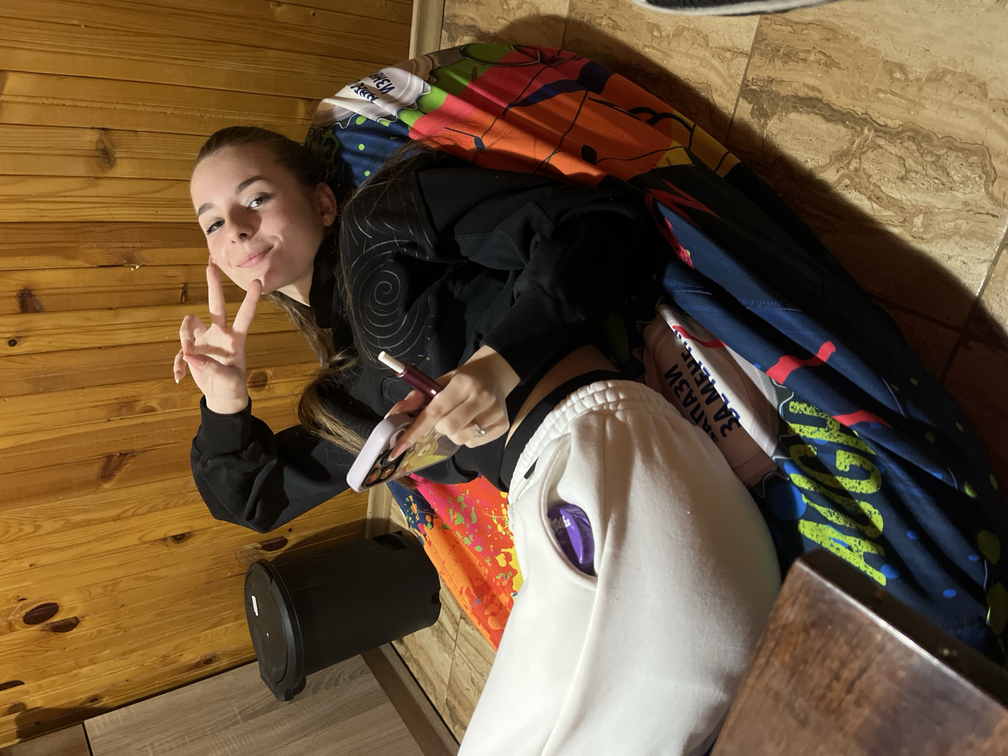 A young woman is lying on a colorful fabric, smiling and holding up a peace sign with her left hand, while holding a phone with a stylus in her right hand. She is wearing a black sweatshirt and white sweatpants. A purple object is visible in the pocket of her sweatpants. To her left is a black trash can. The background consists of wood-paneled walls and tiled floors. There is no information to identify the location in Nova Zagora, Bulgaria.
