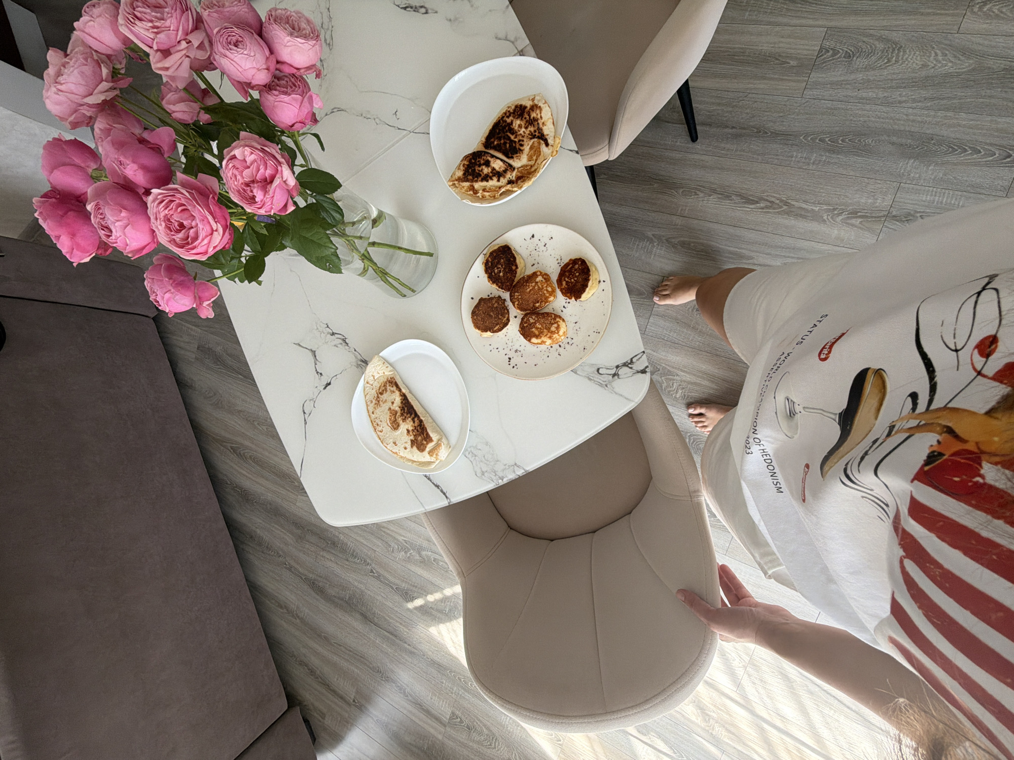 In a room with a marble-patterned table, a vase of pink roses sits next to plates of food. One plate holds several small, golden-brown pancakes, while another contains folded flatbreads. A person's legs and torso are visible in the foreground, wearing a white t-shirt with a graphic print. They are standing near a light-colored chair, with their bare feet on a wooden floor. A large, dark sofa is visible in the background. The scene appears to be a domestic setting.