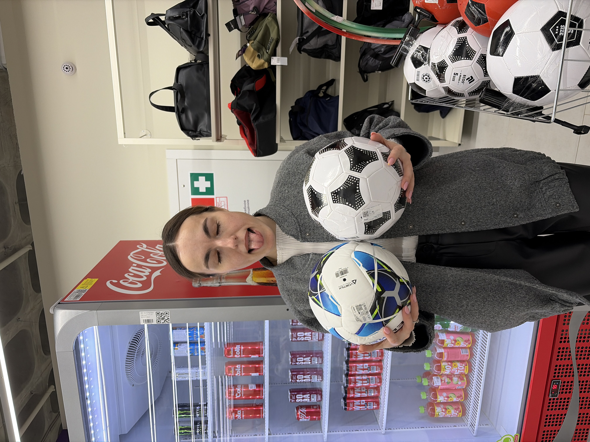 The image shows a woman posing with two soccer balls in front of a Coca-Cola refrigerated display case. The display case contains various beverages, including Coca-Cola products, Monster energy drinks, and other juice beverages. Above the woman and the Coca-Cola cooler are shelves containing various bags. Some soccer balls are displayed above the shelves. The woman is wearing a gray coat and holding a soccer ball in each hand. A first aid kit is located on the wall near the shelving. The text "Izhevsk, Russia" cannot be determined from the image alone.
