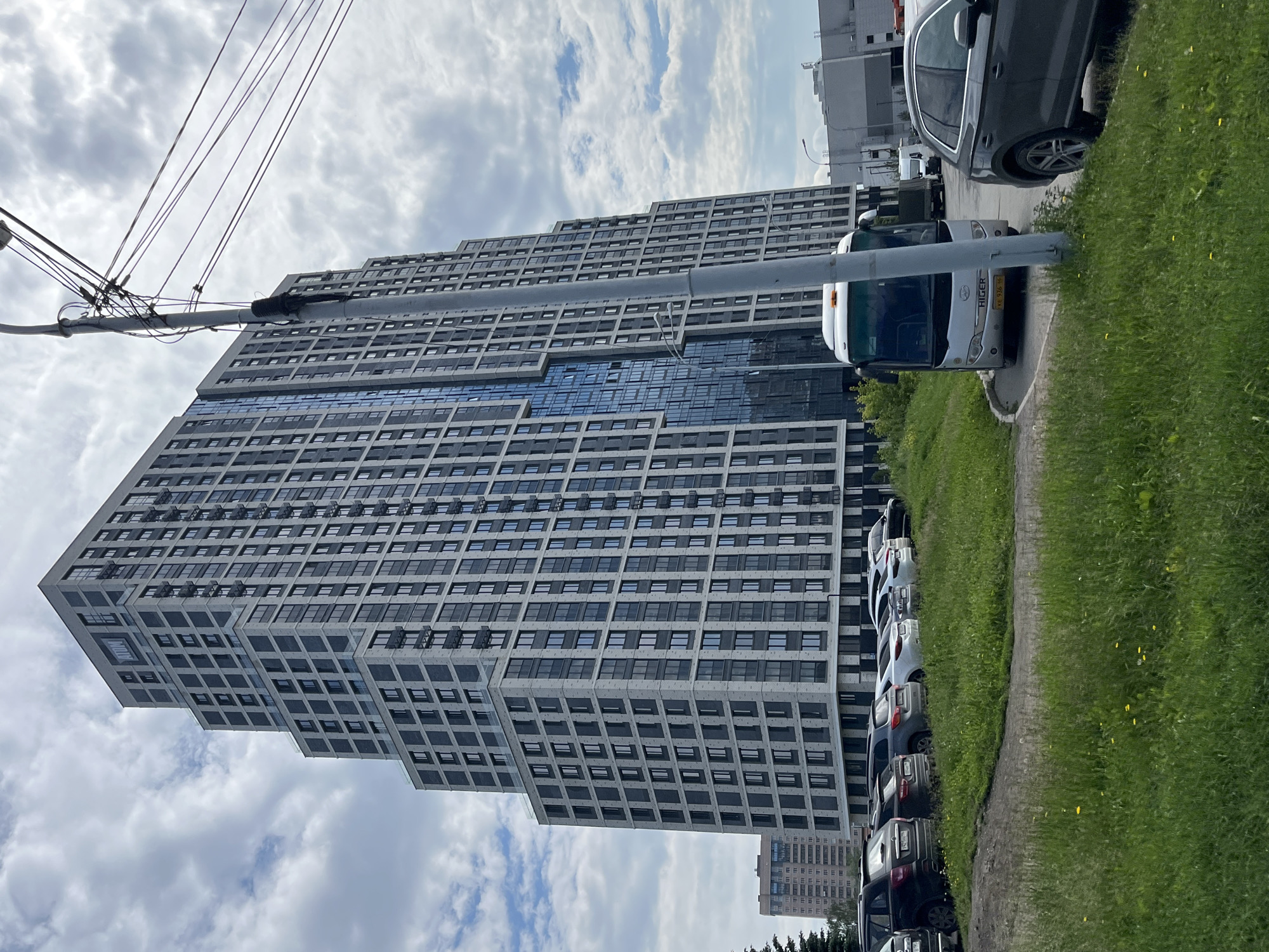 A modern, stepped high-rise apartment building with a facade of light gray brick and dark gray window frames dominates the scene. The building has a prominent glass-covered section. To the left of the high-rise, a slightly shorter, older apartment building with a similar window pattern is visible. In the foreground, a row of parked cars lines a grassy area. A white tour bus is parked on a paved area in front of the high-rise. A utility pole with numerous wires stands in front of the building, and a dark gray SUV is parked on the right side of the image. The sky is overcast with scattered clouds. The location is confirmed to be Yekaterinburg, Russia, specifically the "ATOM" residential complex.