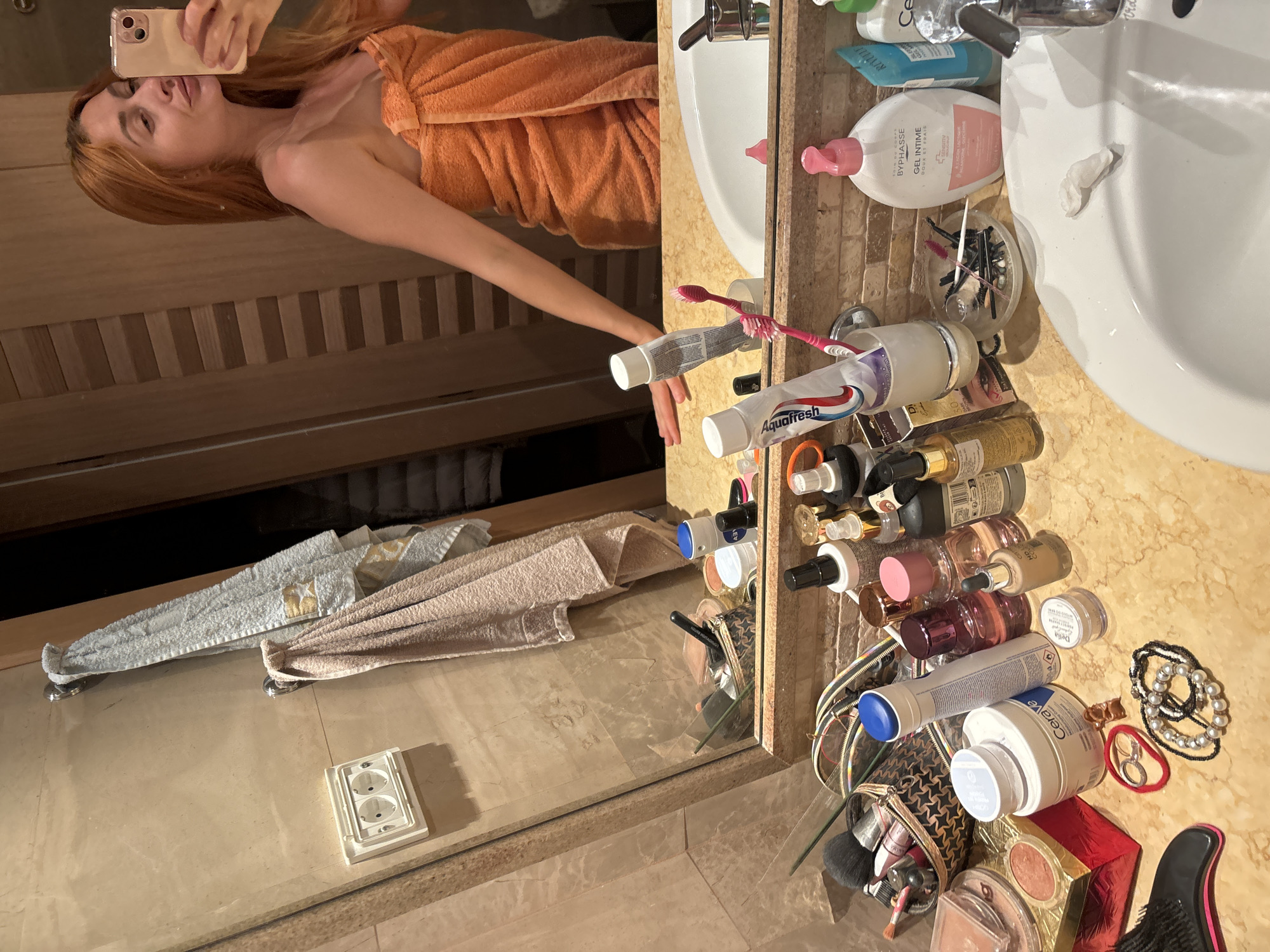 In a bathroom, a woman with long, auburn hair, wrapped in an orange towel, stands in front of a mirror, holding a smartphone and taking a selfie. The bathroom features beige tiled walls and a marble countertop where various toiletries are scattered. Visible items include toothbrushes, toothpaste, makeup products, bottles of lotion and perfume, and jewelry. A sink is present to the right of the countertop. The overall impression is of a personal grooming space.