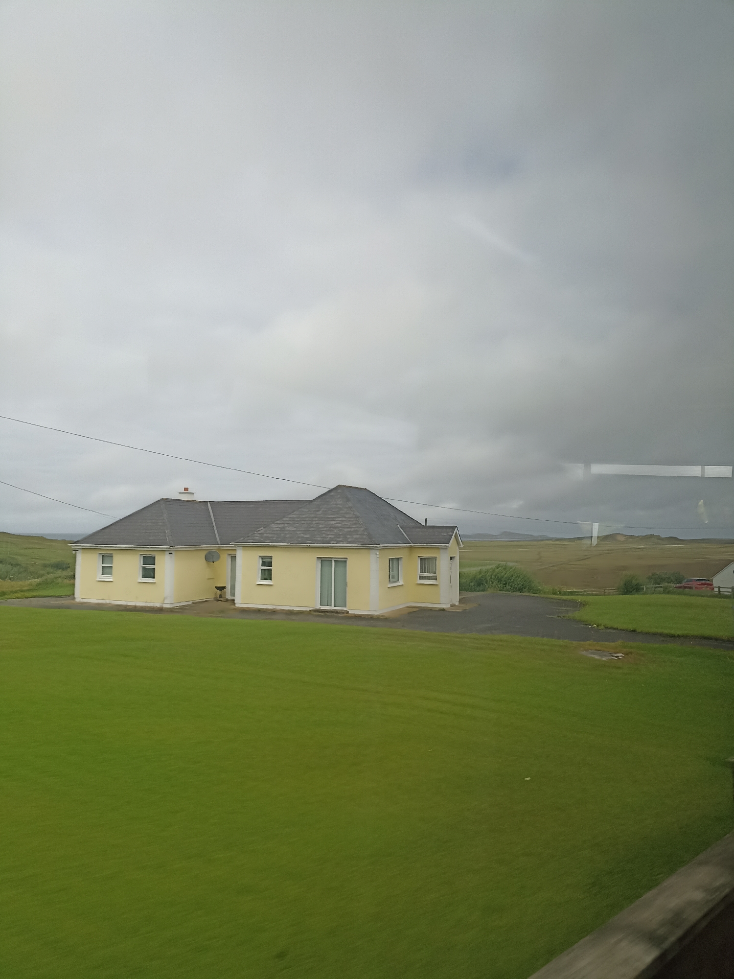 The image shows a yellow, single-story house with a dark grey roof. The house has several windows and a sliding glass door. In front of the house is a large, well-maintained green lawn. A dark grey driveway leads to the house. In the background, there are rolling green hills, and in the distance, a body of water can be seen. The sky is overcast with grey clouds. The scene is set in Ranafast, Ireland, a rural area known for its natural beauty. There are no people or animals visible in the image. The overall impression is one of a peaceful, isolated dwelling in a scenic landscape.