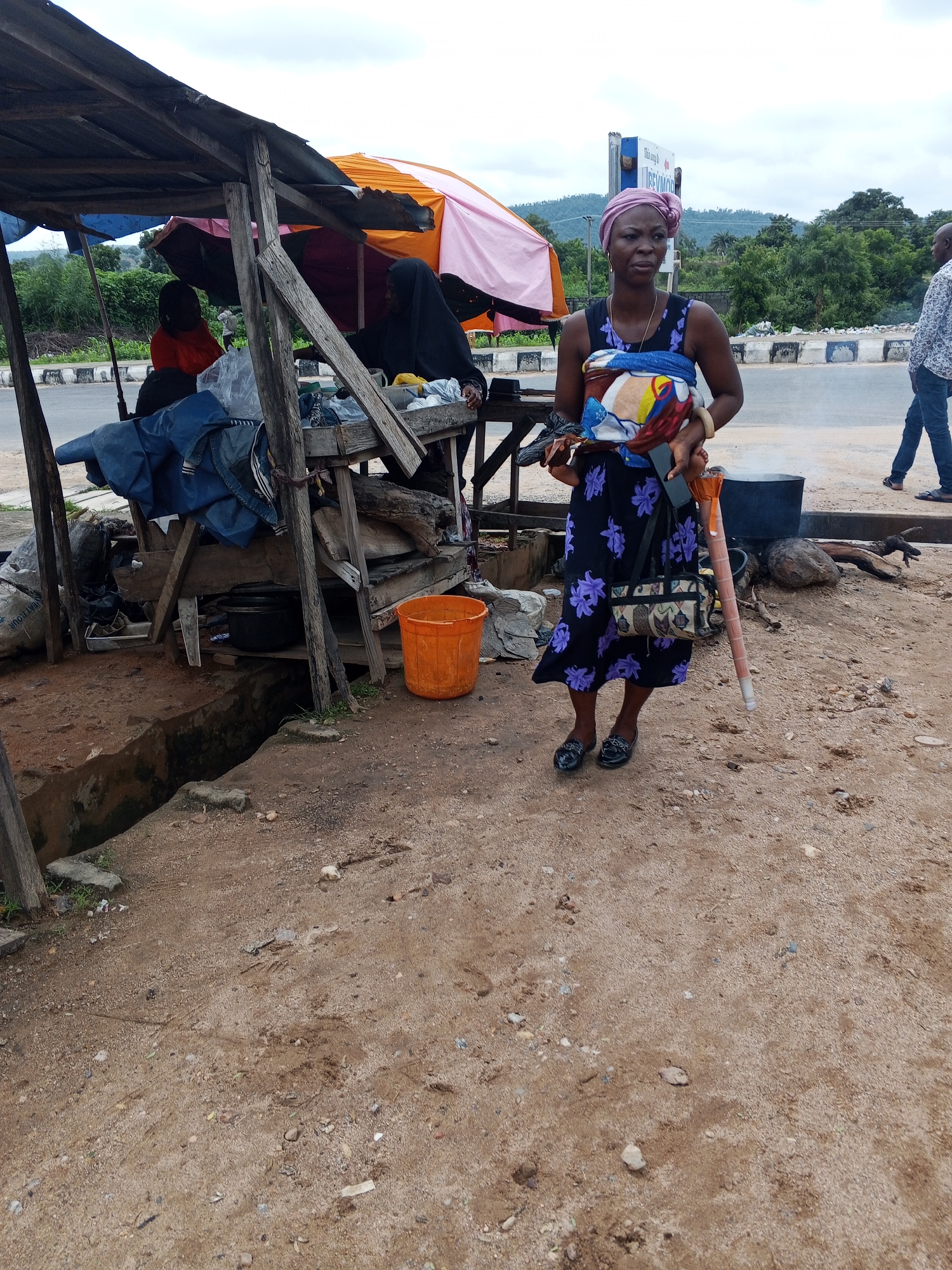 An overhead shot captures a scene in Zango-ndaji, Nigeria, where a woman stands in front of a roadside stall. She is wearing a purple headwrap and a dark floral-patterned dress. She cradles a baby wrapped in colorful fabric. A patterned handbag hangs from her shoulder, and she holds a long, thin object, possibly a cane, in her right hand. The stall, constructed from rough wooden planks and a corrugated metal roof, displays various items, including blue fabric and a black pot. An orange bucket sits on the ground near the stall. The ground is a mixture of dirt and small stones, with a narrow trench running along the side. In the background, a road is visible, with trees and hills beyond. Another person, a man in a patterned shirt and jeans, walks away from the stall. The sky is overcast.