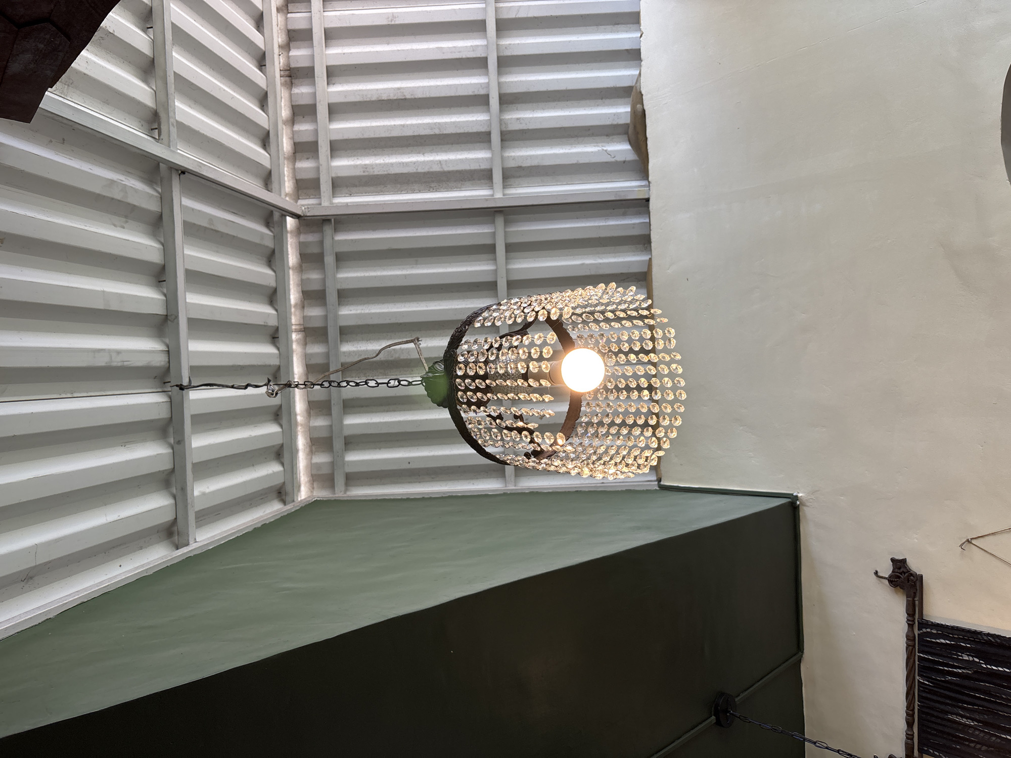 A chandelier with crystal droplets hangs from a chain suspended from a corrugated metal ceiling in Kuta Selatan, Indonesia. The chandelier features a round, dark metal frame from which numerous clear, faceted crystals dangle, reflecting the light of a single incandescent bulb at its center. The ceiling is supported by visible metal beams. To the left, a large section of the wall is painted a muted green, contrasting with a dark, slanted architectural element. Below the chandelier, a cream-colored wall is visible, adorned with a decorative dark wood rod and dark fabric, possibly curtains.