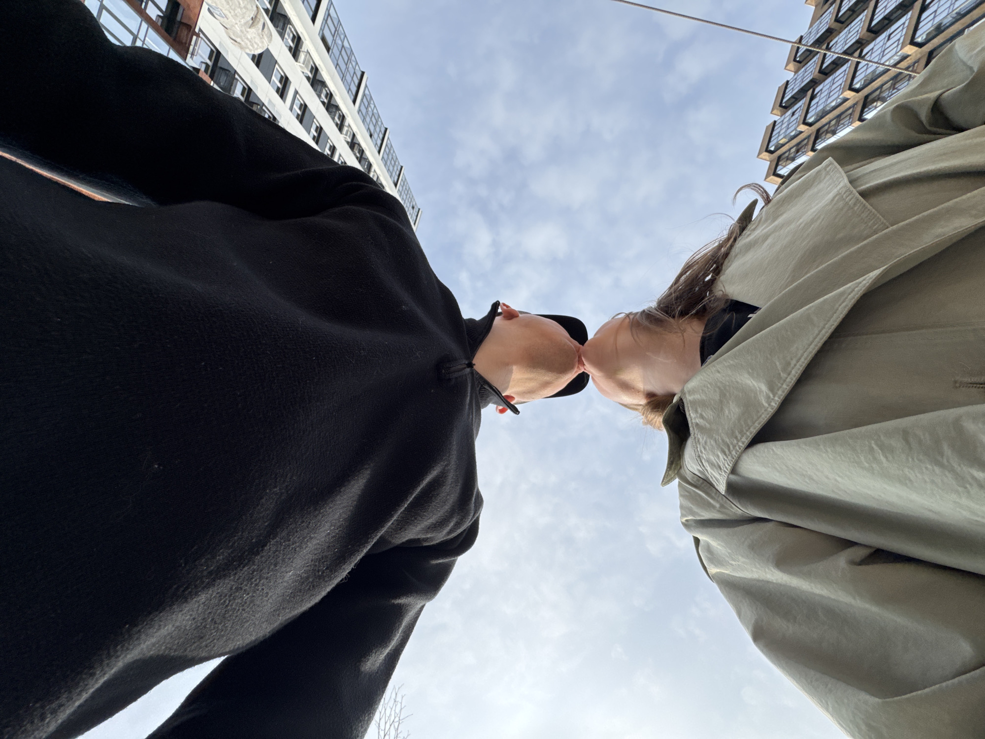 A low-angle shot depicts two individuals kissing, their faces near the center of the frame. The person on the left wears a black hooded fleece, while the person on the right wears an olive-green trench coat. The background shows a cloudy sky and two buildings. The building on the left has a facade of square windows with a white frame, while the building on the right has a facade of square windows with a brown frame. A thin wire stretches from the right side of the image, running across the top of the frame. The location is Perm, Russia.