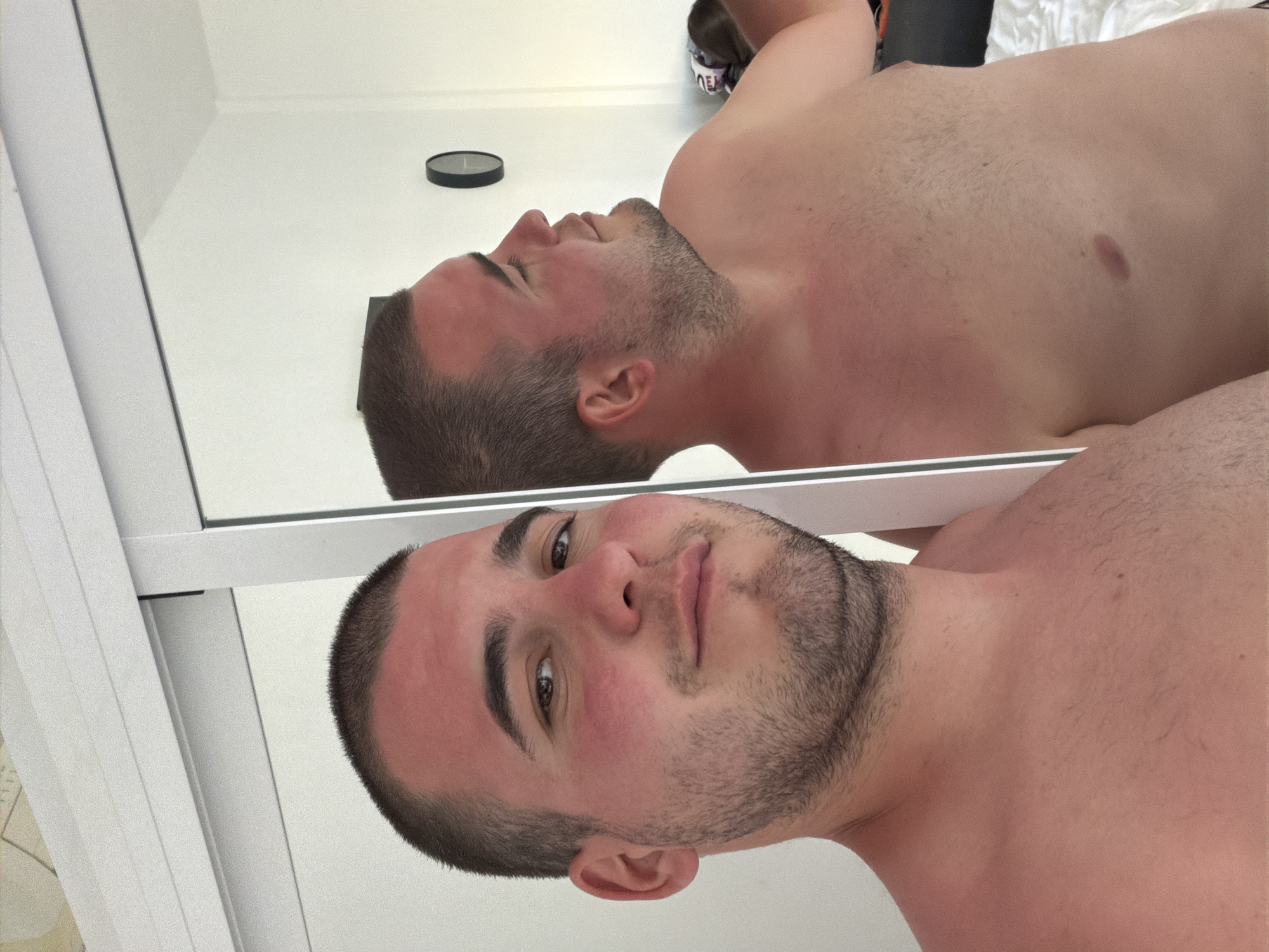 A shirtless man with a short, dark haircut and beard is reflected in a mirror. The man appears to have reddish skin, possibly sunburned. A clock is visible on the wall above the reflected image. The specific location within Arroyo de la Miel-Benalmádena Costa, Spain, cannot be determined from the image alone.