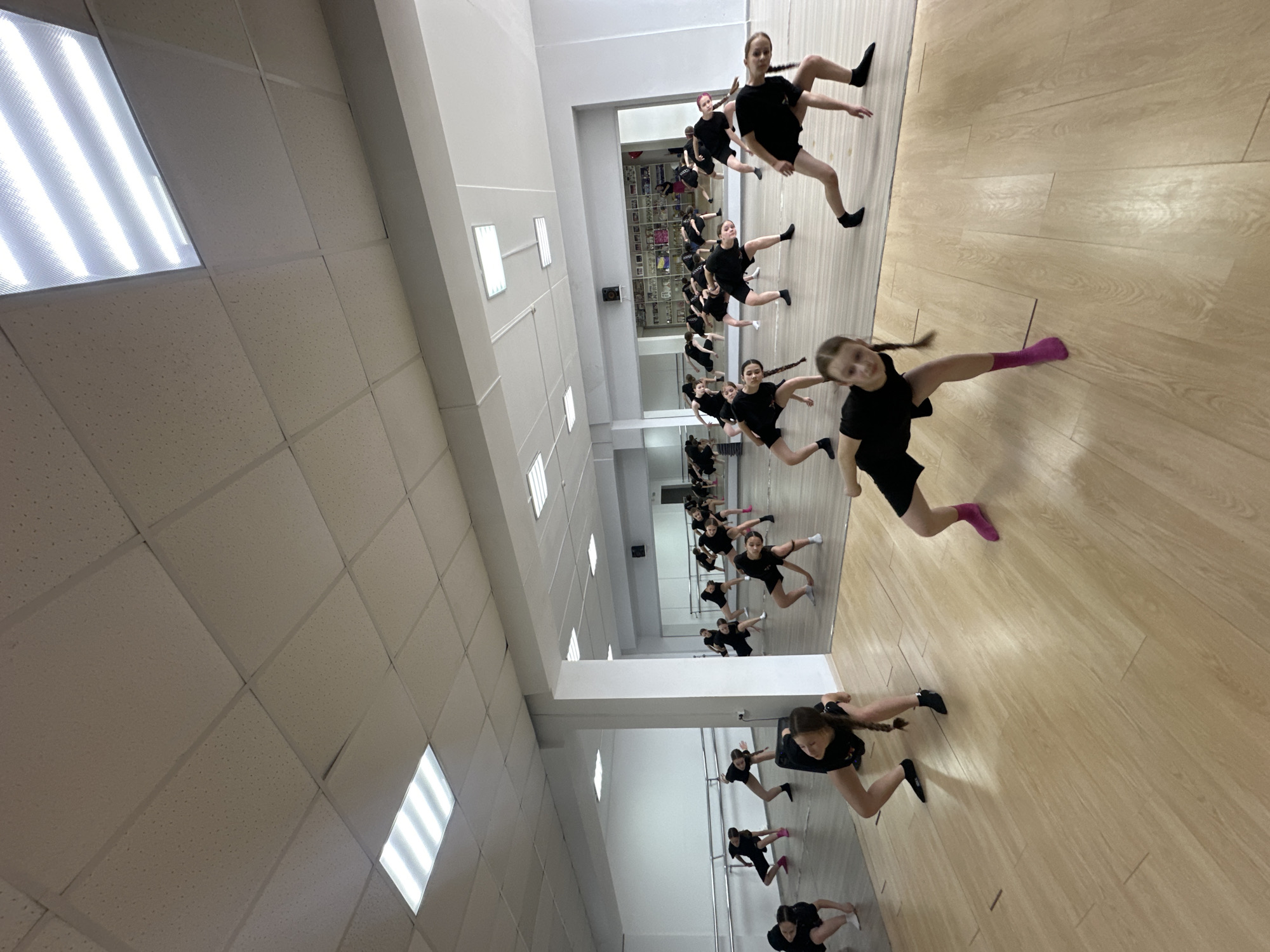 A dynamic scene unfolds within a bright dance studio in Votkinsk, Russia, captured from a low, upward-angled perspective. Numerous young girls, likely pre-teen or early teen, are spread across a light-colored wooden floor, engaged in a dance routine. Each girl wears black athletic attire, consisting of shorts and t-shirts or leotards, and many have their hair in braided pigtails.
The girls are captured mid-movement, striking various dance poses, some appearing to be mid-leap or in deep lunges, showcasing their flexibility and coordination. Their expressions are generally focused, with some displaying slight smiles. Several dancers are visible in the foreground and midground, with many more extending into the background through reflections in a large mirrored wall that spans one side of the room. Along this mirrored wall, white dance barres are visible, providing support for some of the girls.
The room features white walls and a white suspended acoustic tile ceiling, which is fitted with multiple rectangular fluorescent light fixtures that illuminate the space brightly. The overall impression is one of energetic practice or performance. No discernible text is visible within the image.