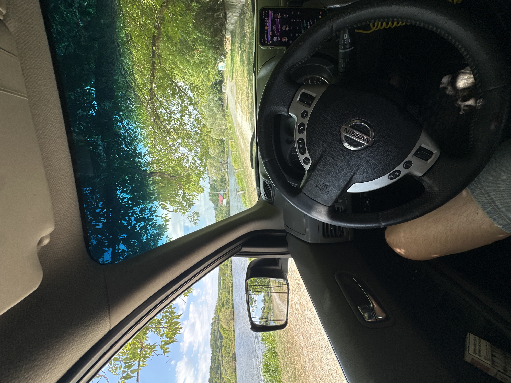 The image is a first-person perspective from inside a Nissan car, parked on a dirt road next to a body of water. A steering wheel with the Nissan logo is prominent in the foreground, and a person's bare leg is visible to the left of the steering wheel. A smartphone is mounted on the dashboard. Through the windshield and side window, a calm body of water, a dirt path, lush green trees, and a clear blue sky with scattered clouds are visible. In the distance, hills and some buildings can be seen. A side mirror on the left shows a reflection of the road and surrounding landscape. The scene appears to be a rural or semi-rural setting, possibly near Makiv, Ukraine, given the context of the location provided. The lighting suggests it is daytime, with sunlight filtering through the trees.
