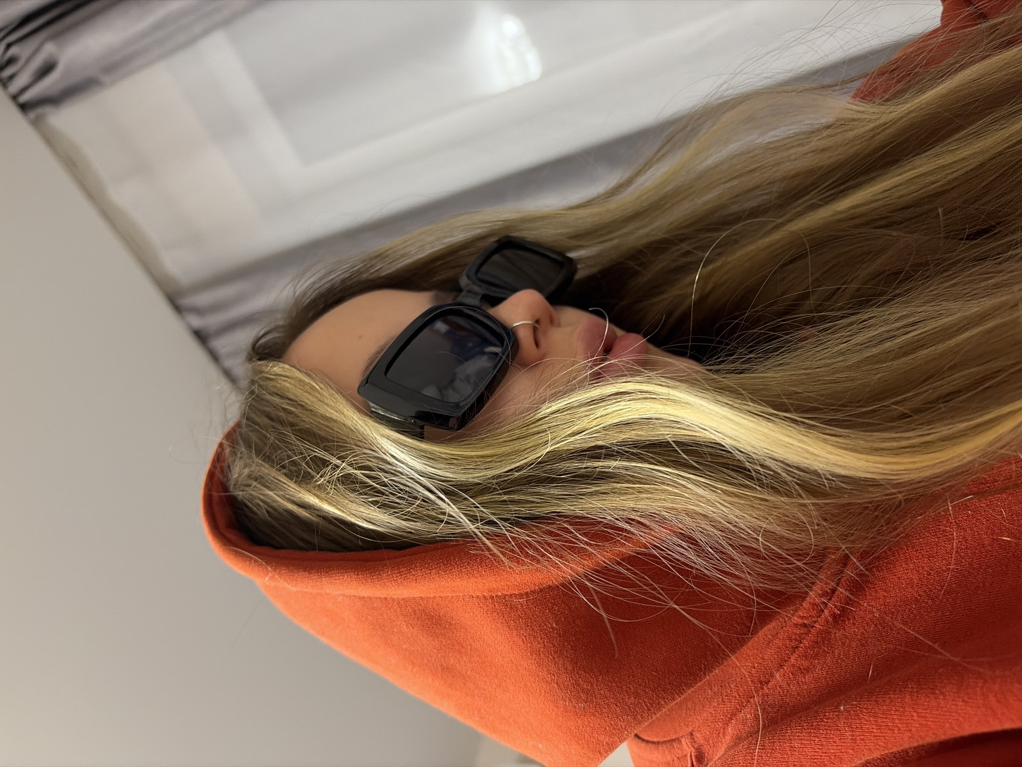 A person wearing an orange hoodie and black sunglasses is lying down. The person has blonde hair and a nose ring. The background is blurry and appears to be a white or light-colored surface. The location in Krasnodar, Russia is not evident from the image.