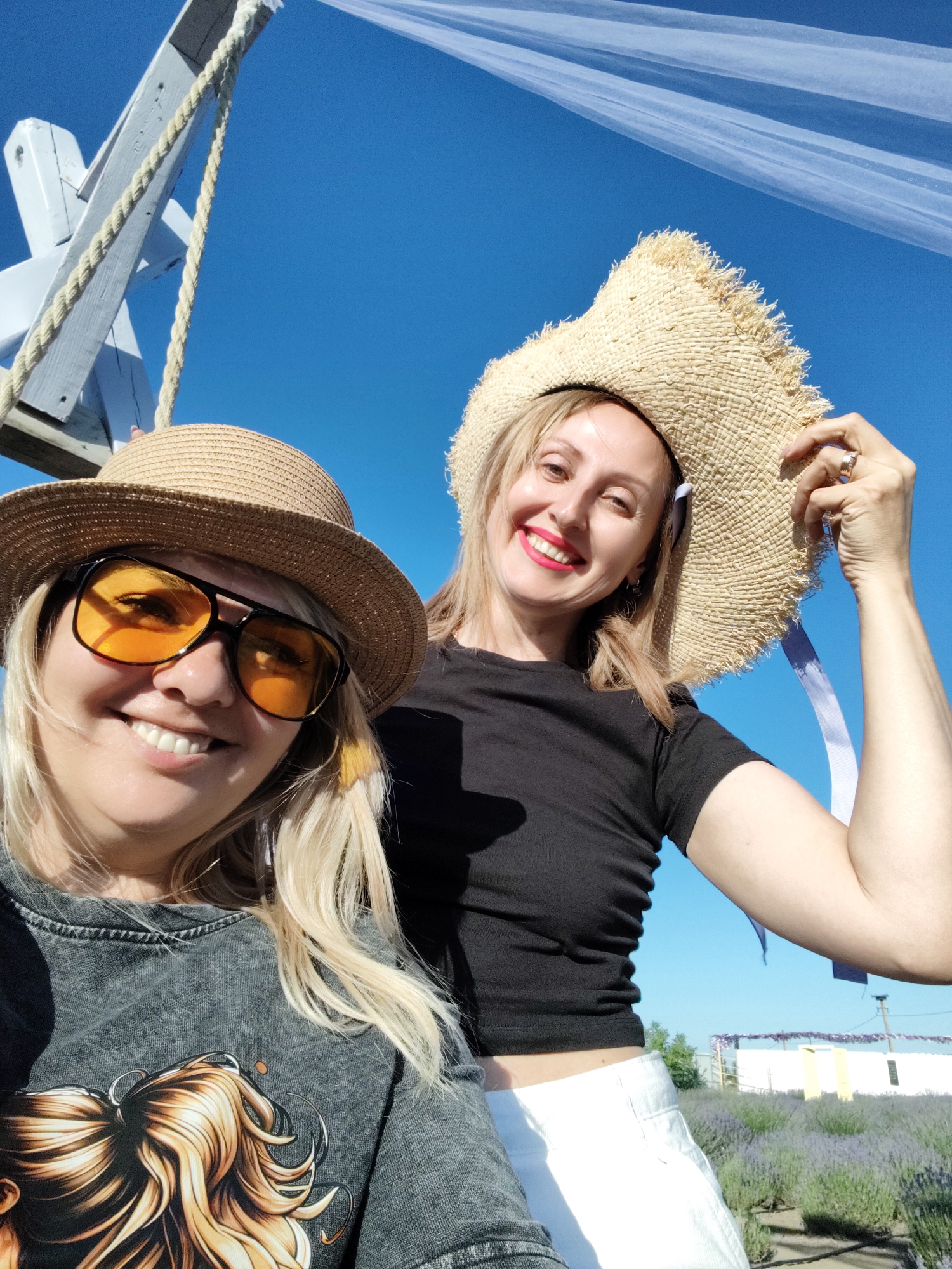 Two women are taking a selfie outdoors on a sunny day. The woman in the foreground is wearing a straw hat with an orange-tinted sunglasses and a dark gray t-shirt with a graphic print. She has blonde hair and is smiling broadly at the camera. The woman in the background is also wearing a straw hat and a black t-shirt. She is holding her hat with her right hand and smiling. Both women appear to be happy and enjoying themselves.
The background shows a clear blue sky and a field of lavender. There is also a white structure with purple flowers hanging from it and a wooden structure with a rope in the upper left corner. The overall scene suggests a warm, pleasant day, possibly in a lavender field or a park. The time of day appears to be late morning or early afternoon, given the bright sunlight and clear sky. There is no visible text in the image.