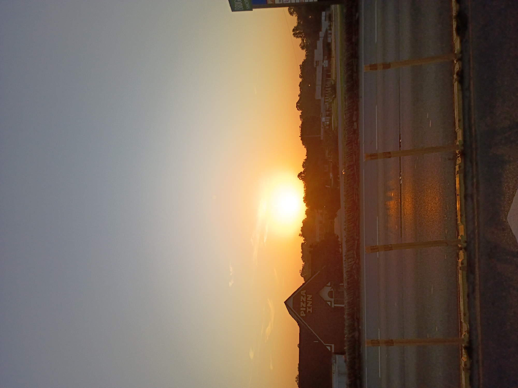 The image depicts a sunset over a road, likely in Frankfort, United States of America. The sun is a bright, yellow orb low on the horizon, casting a warm, orange glow across the sky and illuminating the surrounding trees and buildings. The sky transitions from a pale blue at the top to a vibrant orange and yellow near the sun.
In the foreground, a road stretches horizontally across the frame, with visible lane markings. The road surface appears wet and reflective, suggesting recent rain or dew. Along the side of the road, yellow posts with reflective tape are placed at intervals, likely serving as traffic markers or delineators. A Pizza Inn restaurant building is visible to the left of the road, its roofline sharply defined against the sunset. The building is constructed of brick and has a sign with "PIZZA INN" written vertically on its gable end. Further back, a line of trees and some buildings are silhouetted against the colorful sky.
The overall scene conveys a sense of calm and tranquility, typical of a sunset. The light suggests it is either early morning or late evening. No people are visible in the frame. The visible text includes "PIZZA INN" on the restaurant's sign and partial text on a distant sign that reads "BRIGGS SHOP" and "SHOPPING".