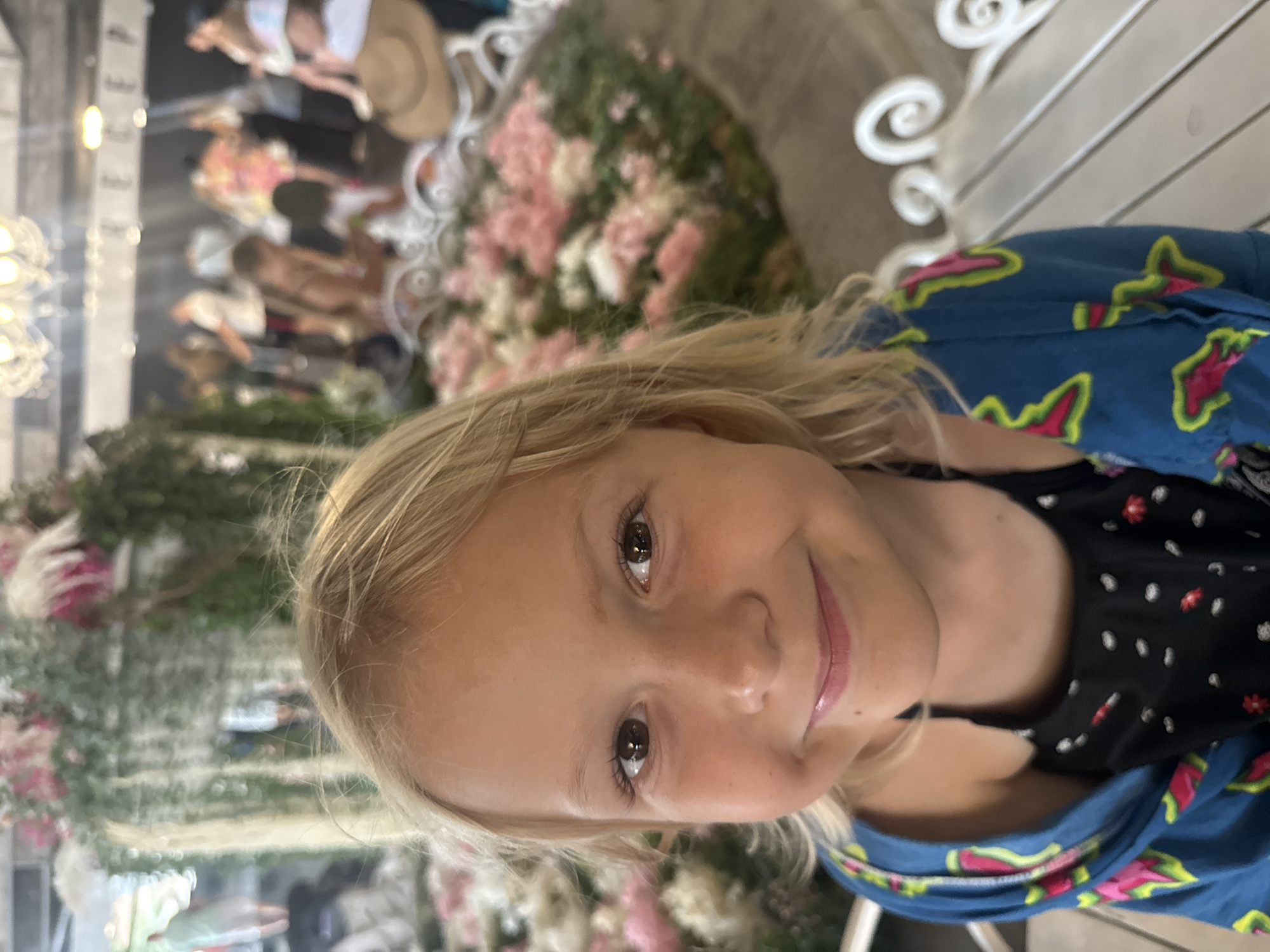 This is a vertical, medium shot of a young girl with blonde hair, looking directly at the camera and smiling. Her eyes are wide and dark, and her lips are slightly parted, revealing a hint of pink lipstick. She is wearing a blue hooded jacket with a colorful pattern of pink and green shapes, and a black top with a white and red floral print underneath. Her hair is a light blonde, with some strands falling across her face.
In the background, out of focus, there are glimpses of other people and floral decorations. The overall impression is a casual and happy scene. The lighting suggests it is daytime. The floral decorations and style of the people in the background could indicate an outdoor event or a decorated indoor space, possibly a venue for a celebration. The hints of people in the background, some appearing to be adults, suggest the girl might be at an event with family or friends. The setting appears to be outdoors or in a well-lit, airy space. There is no visible text in the image.