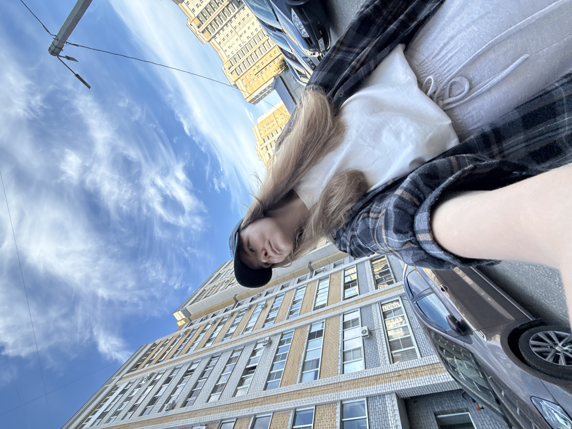 A young woman wearing a dark baseball cap, a white t-shirt, grey sweatpants, and a plaid shirt is standing on a street, taking a selfie with a tall apartment building behind her. The building is light-colored with yellow accents and many windows. Several cars are parked on the street, including a dark-colored sedan in the foreground. The sky above is blue with wispy white clouds. The scene appears to be in a residential area of the city of Vladimir, Russia, given the architectural style of the buildings.