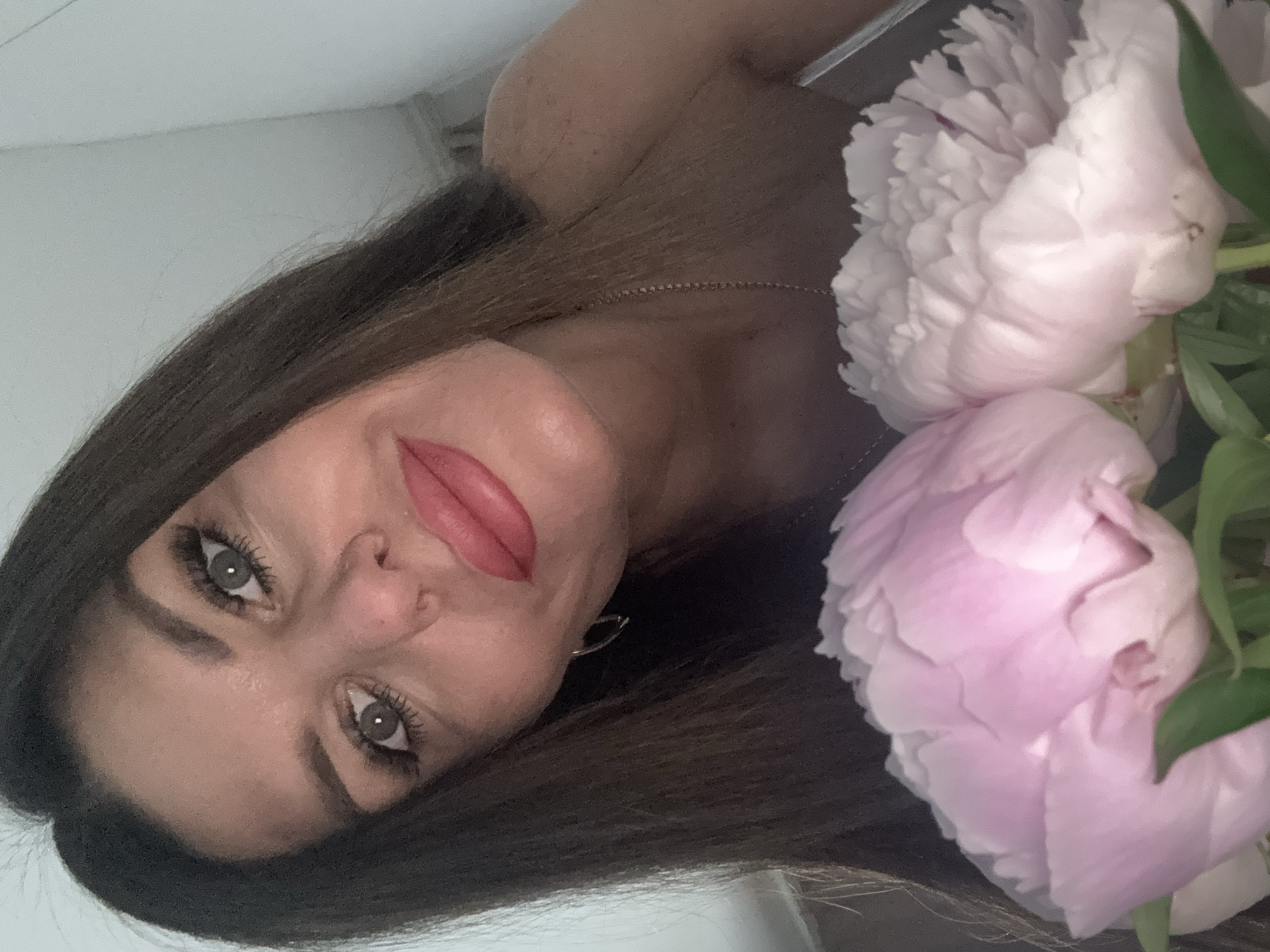 A close-up, high-angle, indoor, selfie-style shot shows a woman with long, dark brown hair and striking green eyes. She is looking directly at the camera with a slight smile. Her lips are painted a vibrant coral-pink, and she wears subtle eye makeup and a small silver hoop earring in her left ear. A delicate silver chain necklace is visible around her neck. In the foreground, obscuring the bottom of the frame, are two large peony flowers, one a soft pink and the other a creamy white, with visible green leaves. The background is a plain, light-colored wall, suggesting an indoor setting. No specific location within the city of Chaikovsky, Russia, can be determined from the image.