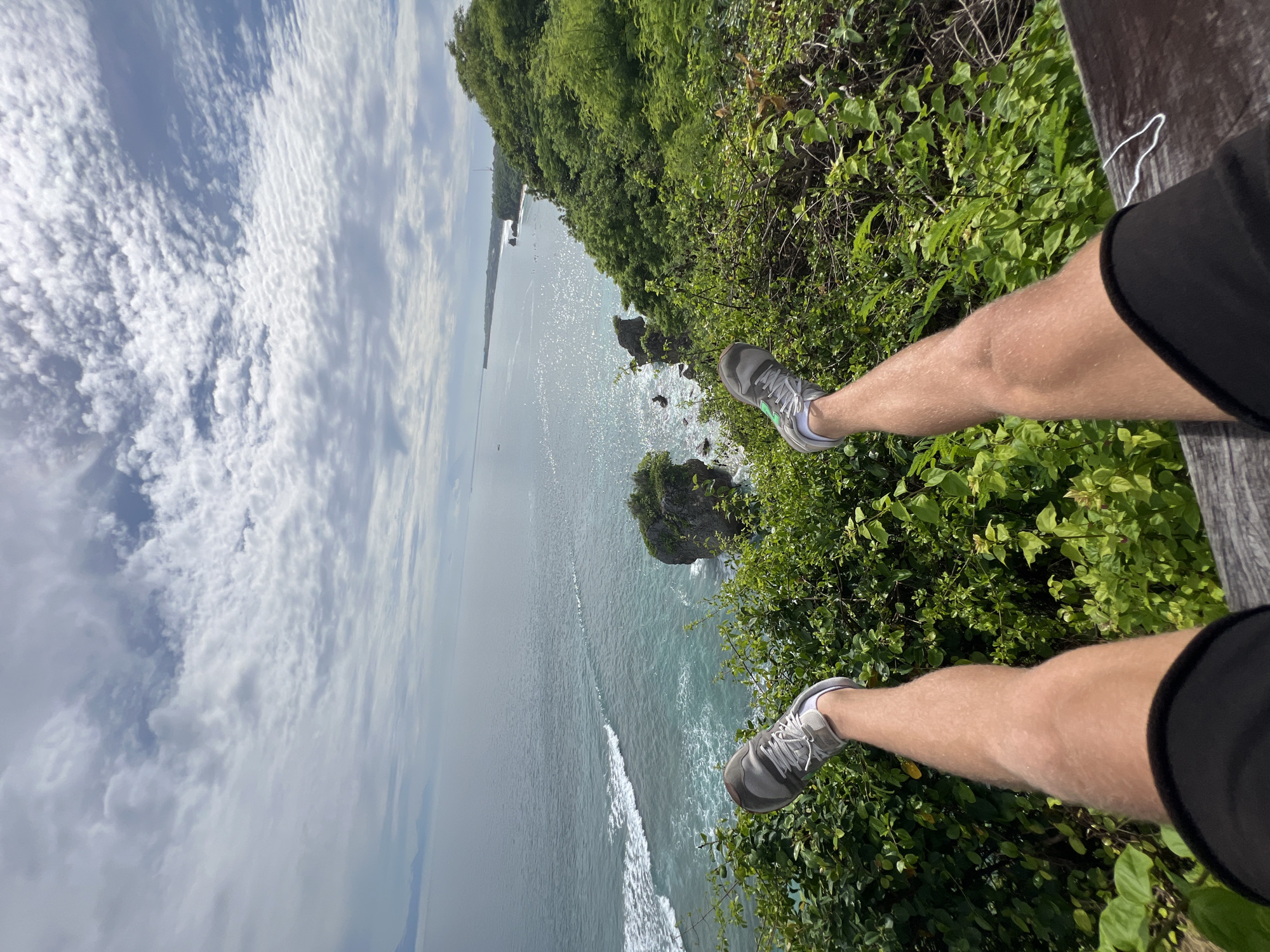 The image shows a pair of legs, dangling over a cliff edge in Kuta Selatan, Indonesia. The person is wearing gray running shoes with white laces, black shorts, and appears to be sitting on a wooden surface. The landscape consists of dense green vegetation covering the cliff face, with views of the ocean and rocky outcrops below. Waves are visible in the water. The sky is partly cloudy.