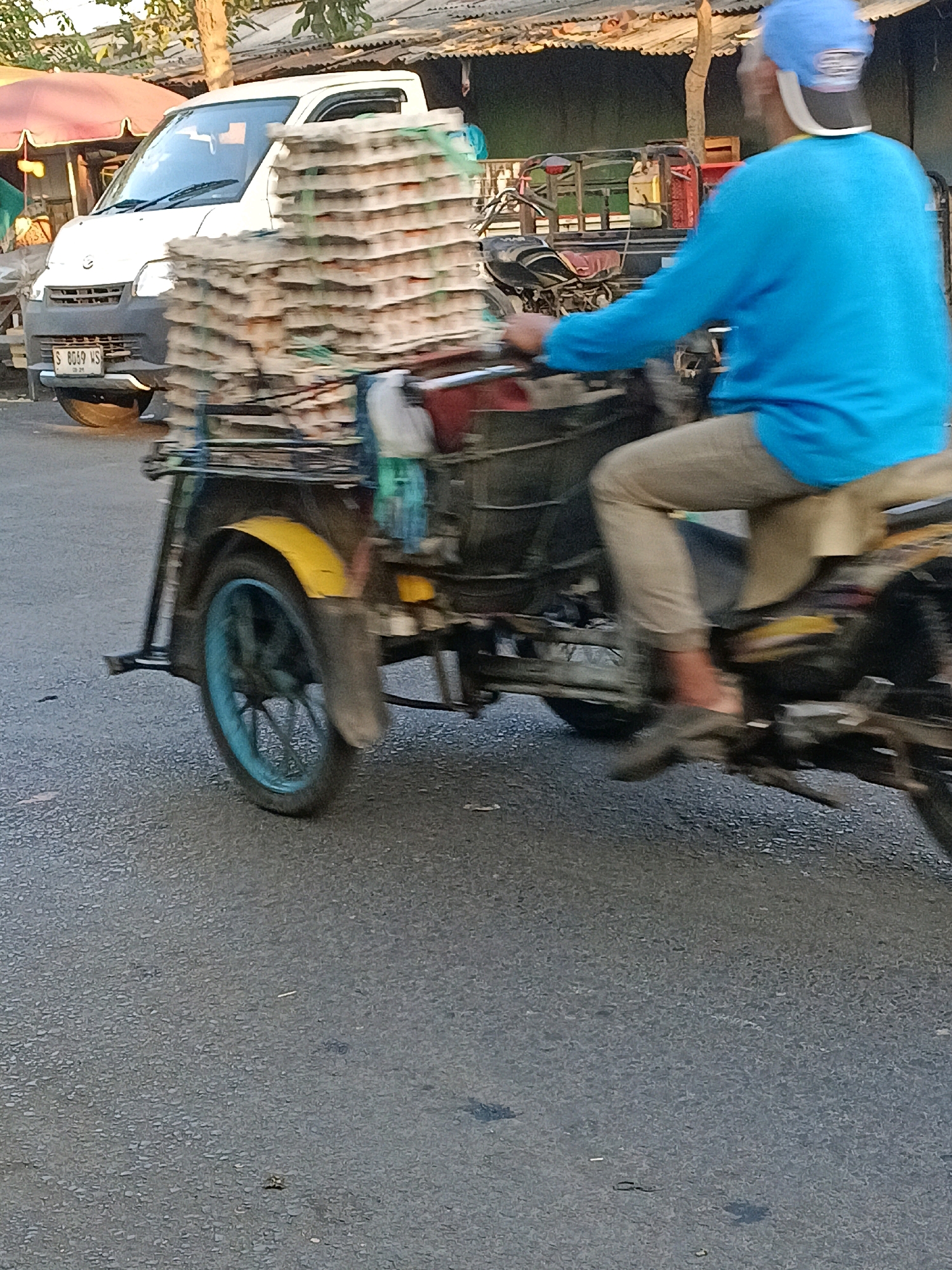 In the city of Wonocolo, Indonesia, a person wearing a blue long-sleeved shirt and tan pants is riding a three-wheeled vehicle. The vehicle is loaded with a tall stack of cardboard egg cartons. The rider is wearing a blue baseball cap. In the background, a white van is parked on the side of a road, and a red and white umbrella is visible. The scene appears to be a street with a rough asphalt surface, with some vegetation and buildings in the background, suggesting a commercial or residential area. The image is slightly blurred, indicating movement.