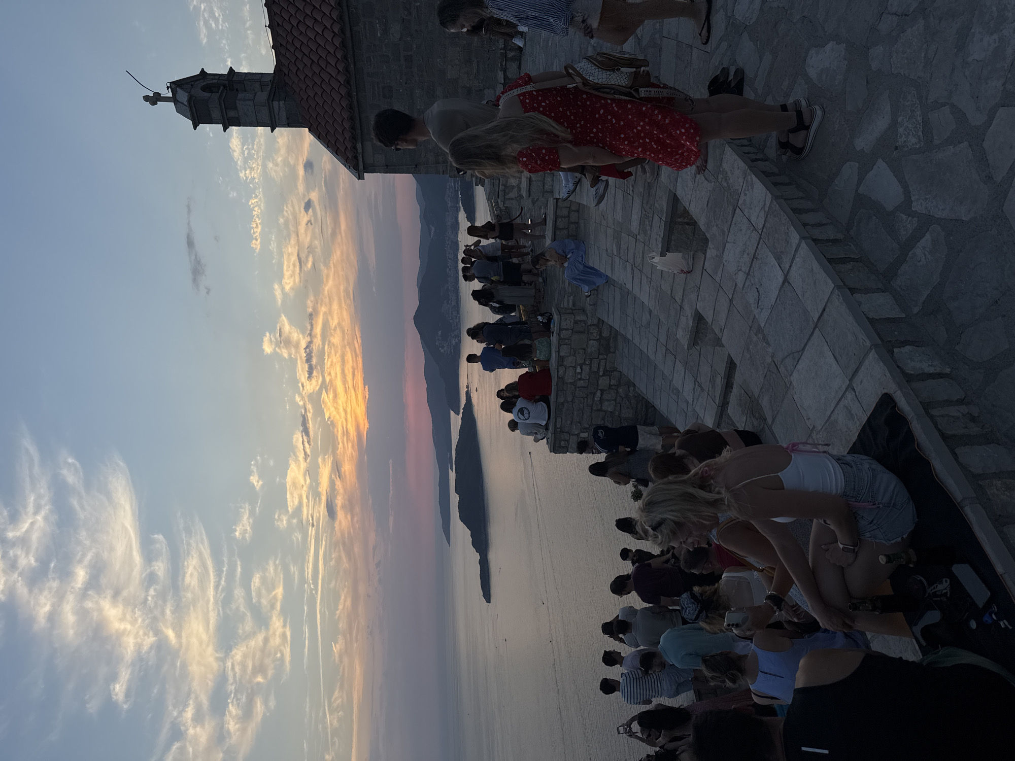 This is a wide shot of a group of people gathered on a stone terrace overlooking the sea at sunset, likely in Sveti Stefan, Montenegro, given the context. A small stone church with a bell tower and a cross on top is visible on the right side of the frame, partially obscured by the people. The sky is filled with a dramatic sunset, with hues of orange, pink, and purple painting the clouds. The sea stretches out to the horizon, with several islands and the coastline visible in the distance. The people are casually dressed, and many are taking photos or videos with their phones. Some are sitting on a low stone wall, while others are standing. The overall atmosphere appears relaxed and celebratory.