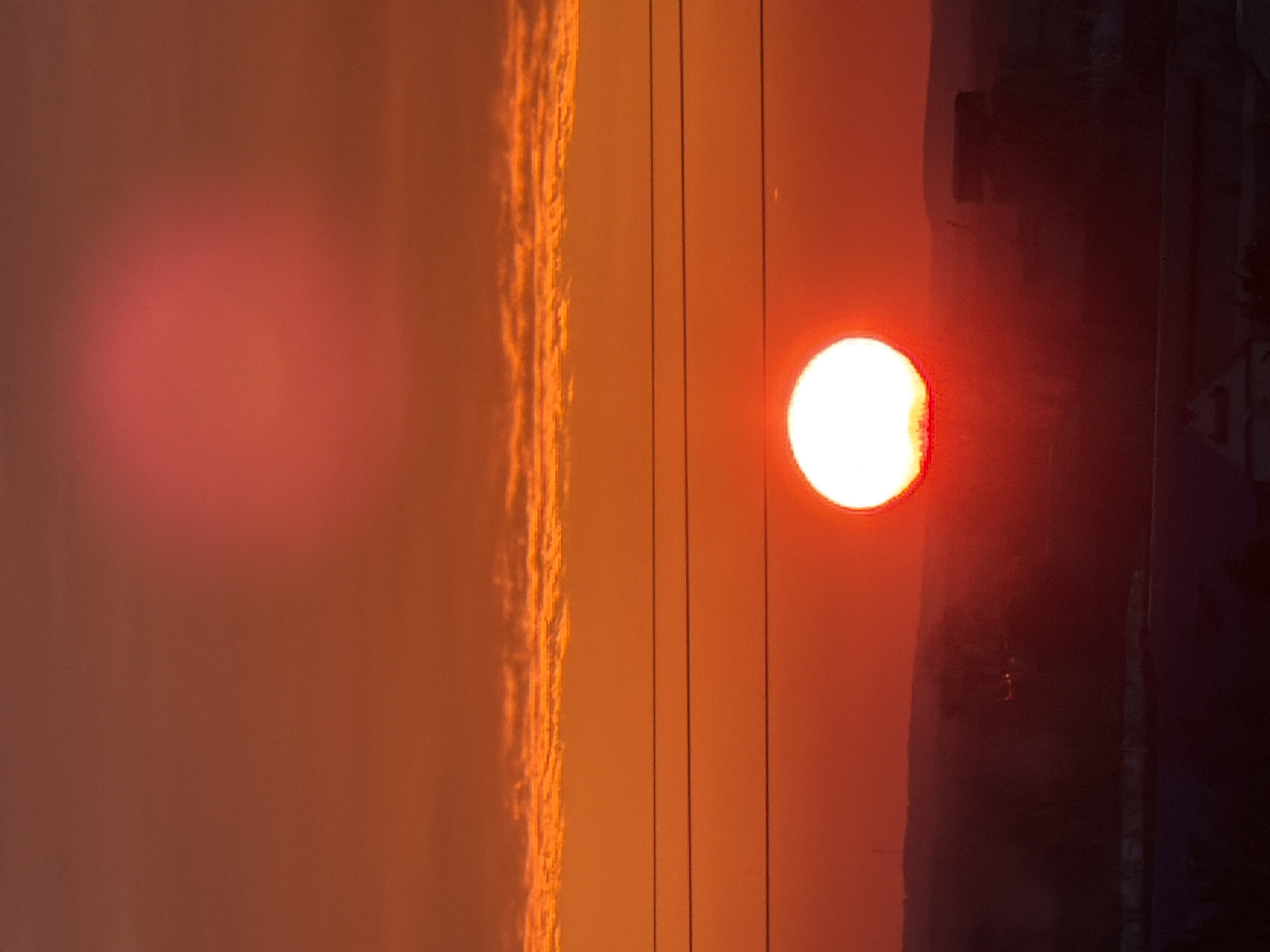 A vertical photograph captures a sunset or sunrise over the city of Chaikovsky, Russia. The sky exhibits a strong color gradient, transitioning from deep red at the lower horizon to vibrant orange and then a softer reddish-orange towards the zenith. A prominent, intensely bright white circular sun disk is positioned in the lower-middle of the frame, its lower portion obscured by the dark silhouette of the foreground. Three dark, thin, parallel utility lines horizontally traverse the image above the sun. A distinct, horizontal band of textured orange clouds is visible in the upper-middle sky. A diffuse red light anomaly is present in the upper sky area. The lower portion of the image, below the sun, reveals dark, indistinct silhouettes of urban structures and distant rolling terrain against the illuminated sky. No individuals, explicit actions, or interactions are discernible within the scene.