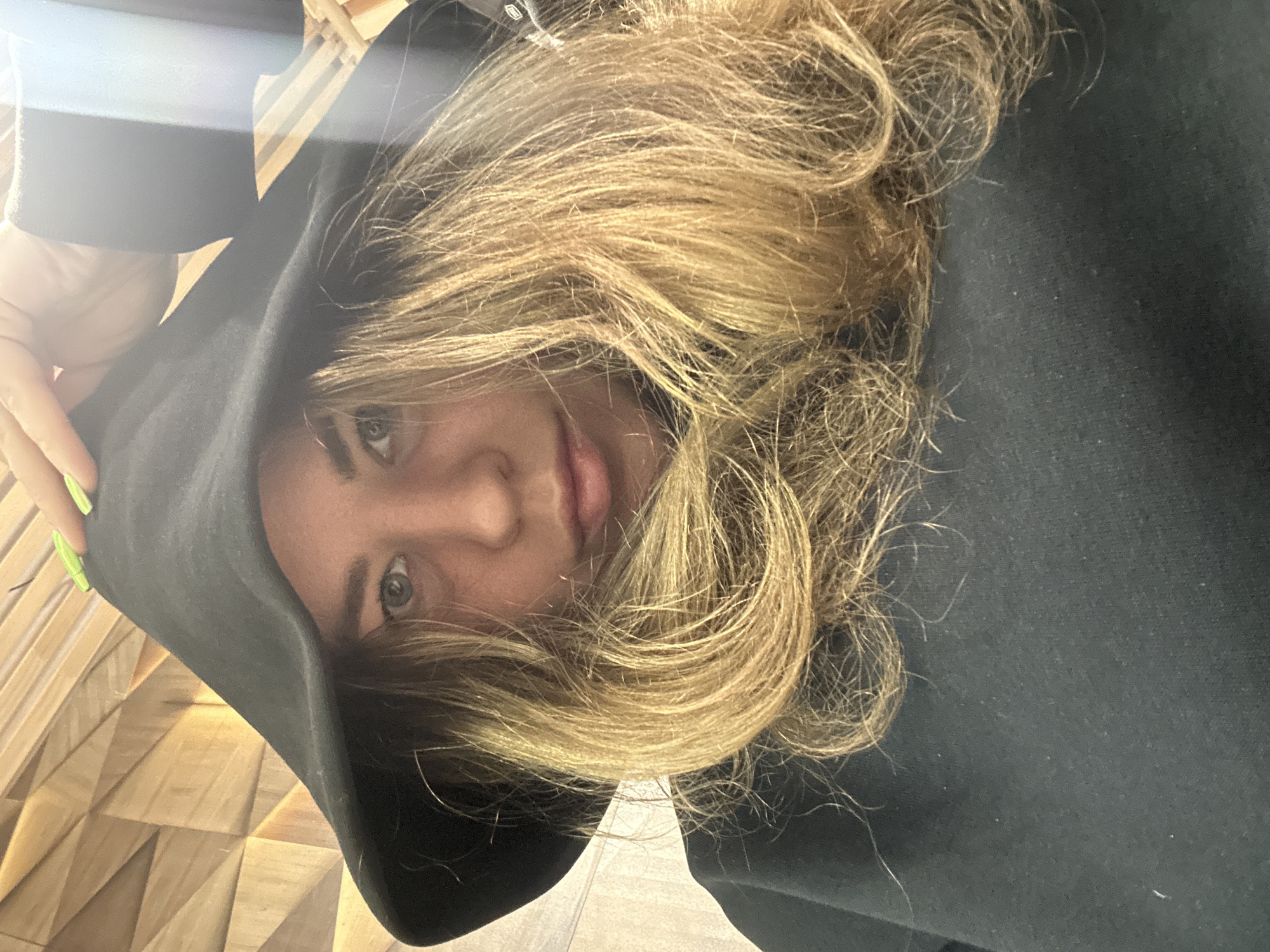 The image is a close-up portrait of a person, rotated 90 degrees counter-clockwise, making the person appear to be looking upwards or sideways in the original orientation.
**Subjects:** The primary subject is a person, likely a young adult, looking directly into the camera. Their face is partially framed by their hair and a dark hood. Their long, wavy, blonde or light brown hair is prominent, appearing somewhat disheveled and flowing around their face. One of their hands, with bright neon green nail polish on the fingertips, is visible on the upper left, holding the edge of the dark hooded garment.
**Objects:** The main object is the dark-colored (likely black or very dark grey) hooded sweatshirt or jacket worn by the person.
**Scene and Setting:** The setting appears to be indoors. A distinctive feature in the background on the left side of the image (when oriented upright) is a textured wall with a geometric, three-dimensional pattern, possibly made of wood or a wood-like material. The rest of the background is obscured by the person's clothing and hair.
**Activity or event taking place:** The person is posing for a selfie or a casual portrait, looking directly at the camera.
**Notable details:** The bright green nail polish provides a pop of color against the dark clothing and light hair. The lighting seems to be soft and even, possibly indicating indoor light or diffused natural light. The person has a neutral to slightly expressive gaze.
**Visible text:** There is no visible text in the image.