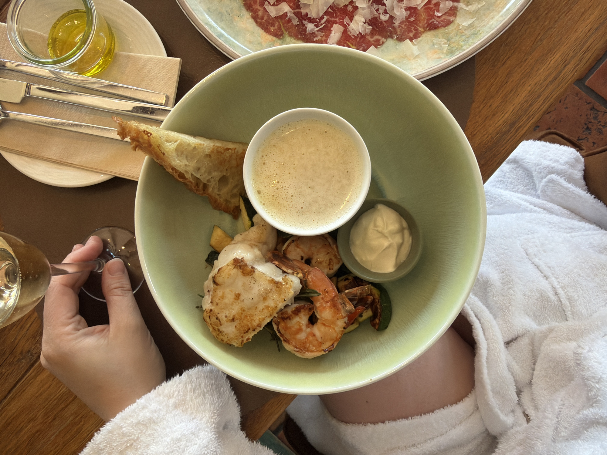 A first-person overhead shot depicts a person in a white robe seated at a wooden table, presumably in Bergisch Gladbach, Germany. The person is holding a wine glass in their left hand. On the table is a light green bowl containing a piece of toasted bread, shrimp, vegetables, and two small bowls filled with sauce. A white plate with cutlery and a jar of yellow liquid is in the upper left corner. A plate of sliced meat topped with shaved cheese is also on the table, out of focus. The focus is on the bowl of food.