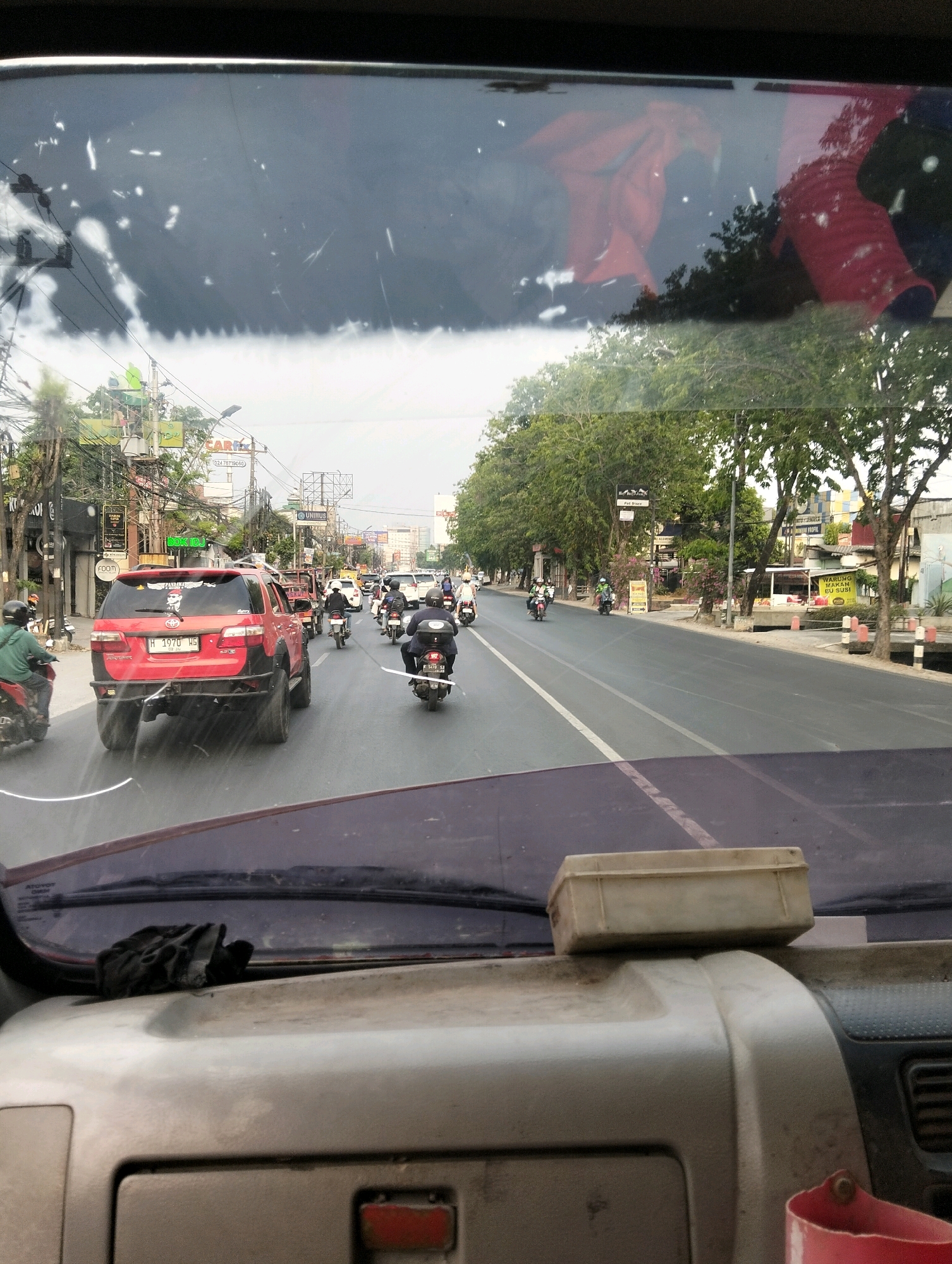 The image was taken from inside a vehicle, looking through the windshield at a busy street in Semarang, Indonesia. The windshield is dirty and reflects parts of the vehicle's interior, including a red cloth object and a white box. The street is lined with trees and buildings, and there is significant traffic, predominantly motorcycles. Several motorcycles are visible, with riders wearing helmets. A red SUV with off-road modifications is in the foreground, also with a sticker on the rear window. Billboards and street signs are also present. The overall impression is of a typical urban environment in Southeast Asia.