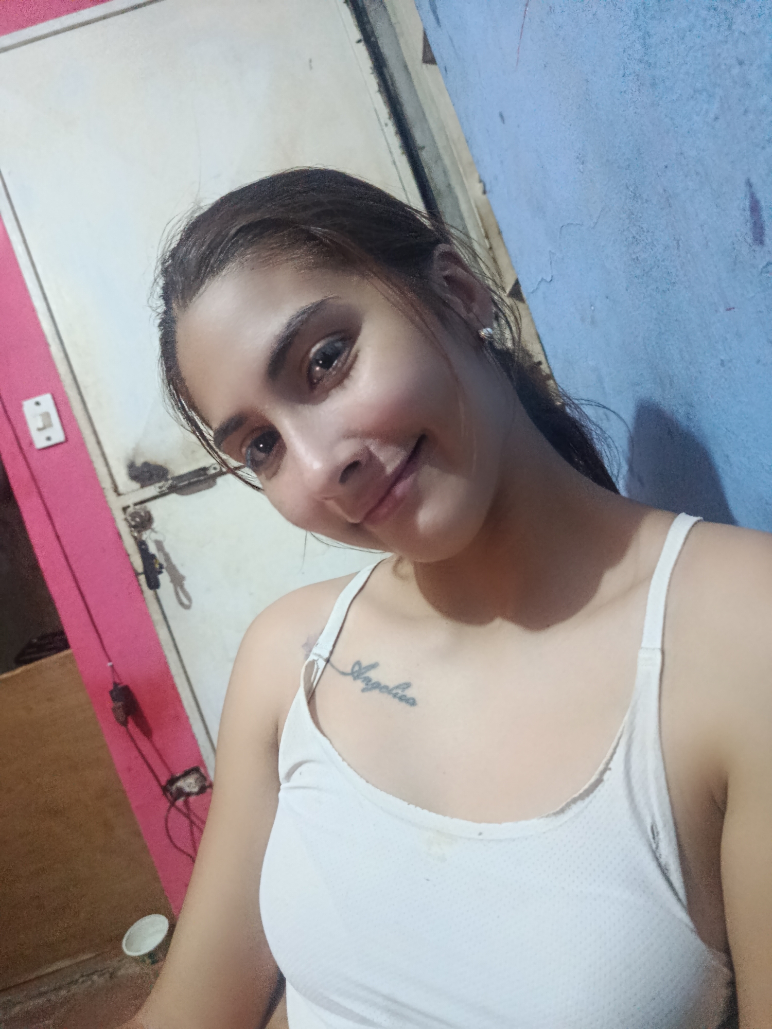 A person is taking a selfie indoors. The person has fair skin, dark hair pulled back, and is wearing a white tank top. A tattoo is visible on the upper chest. The background consists of a pink and white door, keys hanging, a light switch, and a blue wall. The image does not contain evidence to confirm its location in Juan Griego, Venezuela.