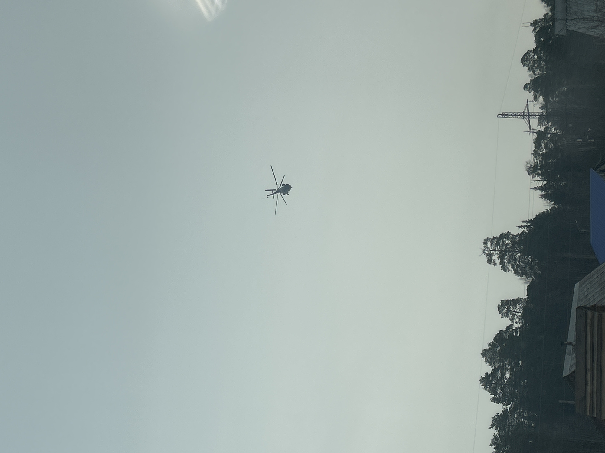 The image depicts a helicopter in flight against a pale, overcast sky. The surrounding environment includes a treeline, a power transmission tower, and a structure appearing to be a building or dwelling. The location is Chaikovsky, Russia. The helicopter is the central subject. The lack of detail prevents further analysis.