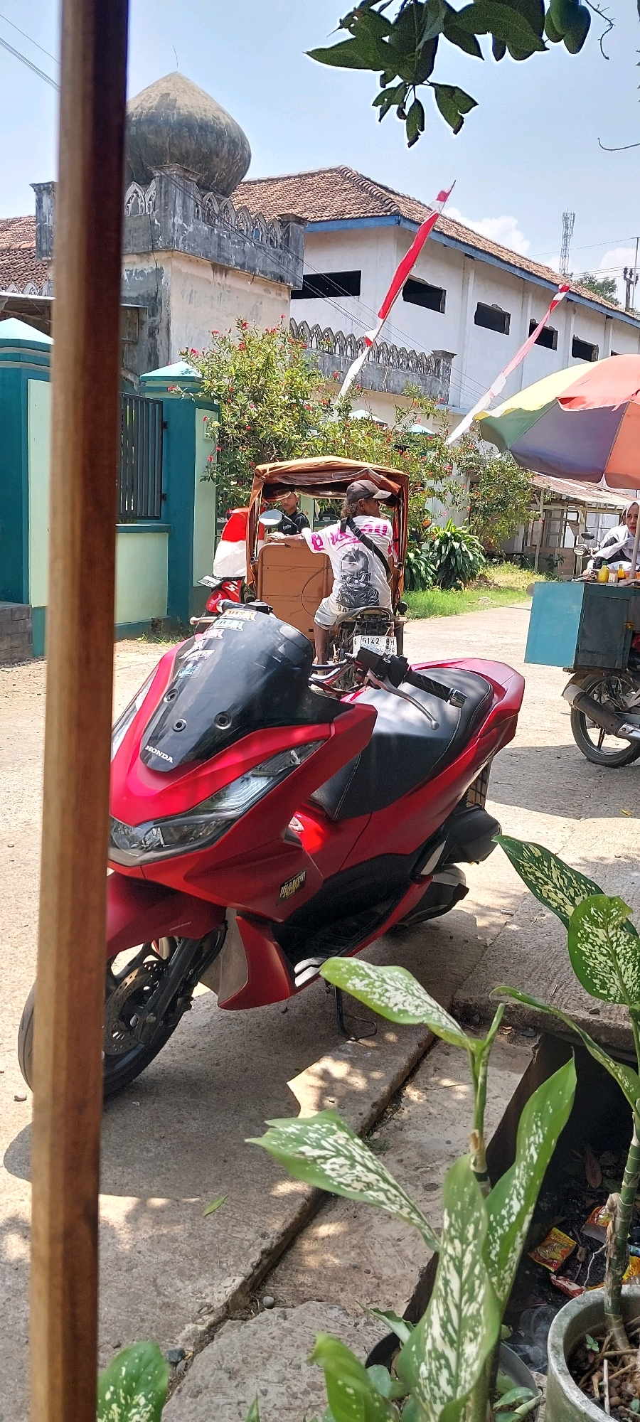 A low-angle, vertical shot captures a red Honda PCX scooter parked on a concrete surface in Keling, Indonesia. The scooter faces right, its front wheel angled slightly towards the viewer. Behind it, a tricycle with a person seated in the driver's seat and another passenger is visible. In the background, a building with a dome structure and red and white flags suggests a cultural or religious site. Trees with lush green foliage and a colorful umbrella add to the outdoor setting. The ground is a mix of concrete and dirt, with some plants in pots to the right of the frame. The sky is bright and clear, indicating a sunny day.