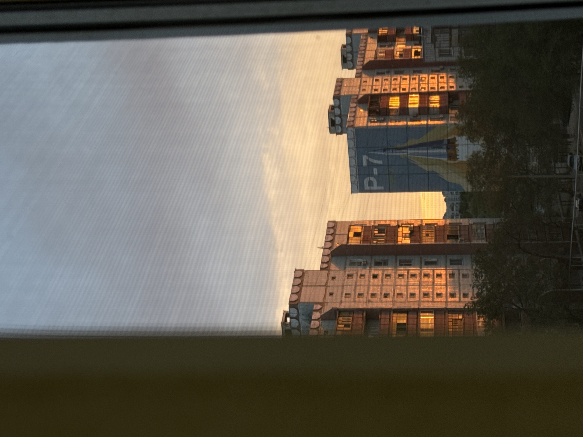 This media file shows a view through a window, likely from an interior space, looking out at an urban scene during sunset. The primary subjects are two apartment buildings, with one prominently featuring a large blue banner with white text that reads "P-7". The buildings are constructed with concrete and have numerous windows, many of which are illuminated by the warm glow of the setting sun. The sky is a soft gradient of pale orange and yellow, indicating it is either sunrise or sunset.
A large tree with dark green foliage partially obscures the right side of the buildings, suggesting an outdoor environment. The overall impression is of a residential area in a city, potentially in a region experiencing twilight. The image is oriented vertically, rotated 90 degrees clockwise. The foreground is dominated by the window frame and a blurred, dark area, likely the interior space from which the photo was taken. The lighting and atmospheric conditions suggest a calm, perhaps reflective, moment at the end of the day. No people are visible in the frame. The provided location context, Baikonur, Kazakhstan, adds a layer of specificity to the setting.