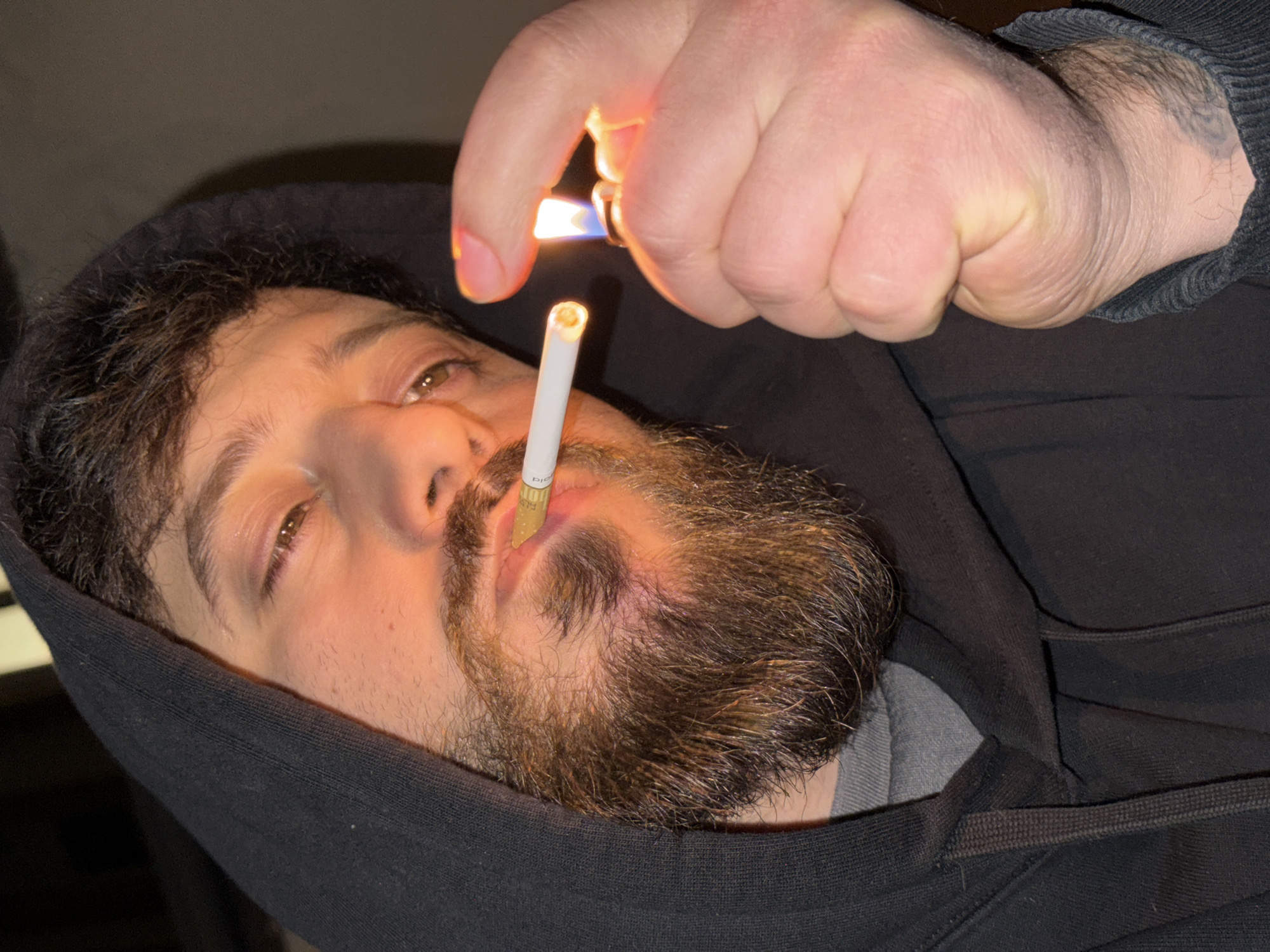 A close-up portrait depicts a male individual in Strasbourg, France. The subject, appearing to be in his twenties or thirties, has dark, curly hair visible beneath a black hooded sweatshirt and a full, dark brown beard and mustache. His light brown eyes are directed towards the viewer. A white cigarette with a light brown filter, partially showing "FILT-" lettering, is held between his lips. His right hand, positioned to the right of his face, holds a metallic lighter producing a prominent blue and yellow flame, actively igniting the cigarette tip. The fingernail of his right index finger is painted or stained orange/peach. A faint tattoo is partially visible on his right wrist, beneath the cuff of his black hoodie. The background is dark and indistinct, with localized artificial light from the lighter illuminating the subject's face, beard, and the immediate foreground.