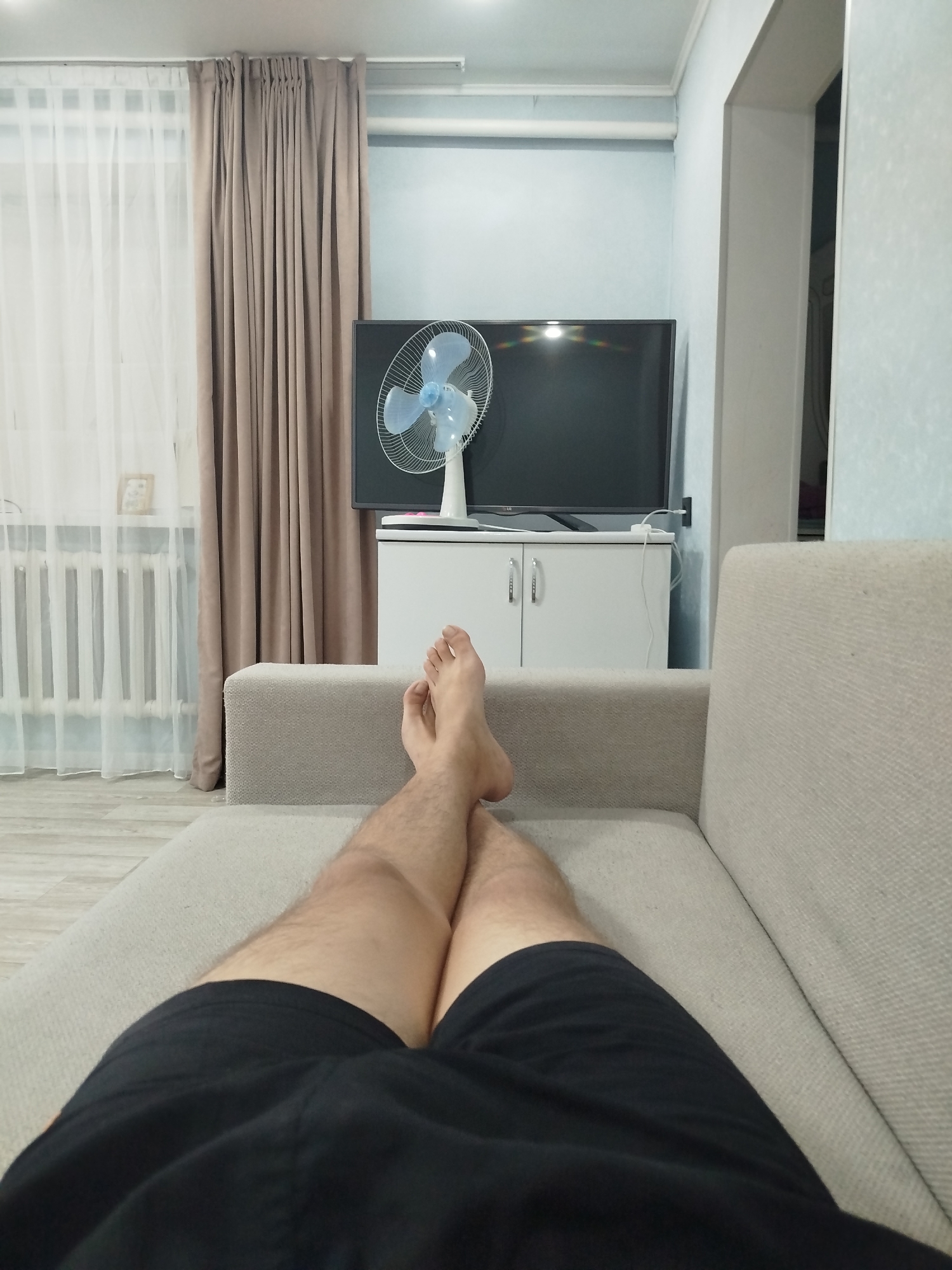 This is a first-person perspective, point-of-view shot taken from a couch in what appears to be a residential living room in Ürjar, Kazakhstan. The scene is brightly lit, likely by indoor lighting, and depicts a relaxed, casual atmosphere.
In the foreground, the subject's legs and feet are visible, resting on the couch. The subject is wearing black shorts, and their bare feet are crossed at the ankles. The couch itself is a light beige or cream color, upholstered in a textured fabric.
The middle ground of the image reveals a white cabinet with two doors, topped by a black flat-screen television. A white fan with blue blades stands on the cabinet to the left of the television. Behind the cabinet and television, the wall is painted a light blue.
To the left of the scene, sheer white curtains are drawn across a window, and behind them, heavy brown curtains are partially visible. A white radiator can be seen beneath the window. The flooring appears to be light-colored laminate or wood. The room seems tidy and well-maintained.