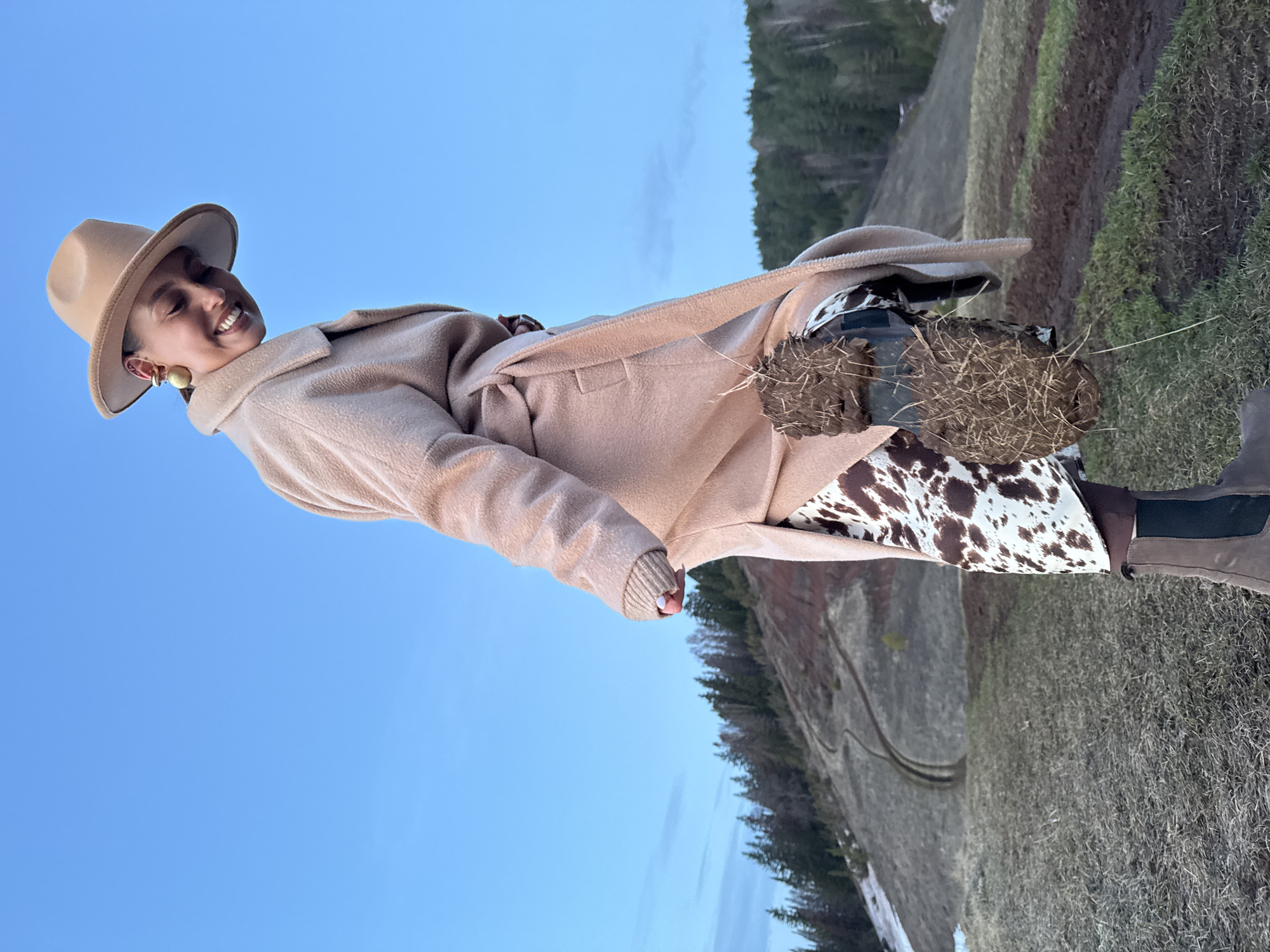A woman is posing for a photograph in an outdoor, rural setting in Ольховка, Russia. She is wearing a tan fedora, a tan coat, a cow-print dress, and brown boots. She is smiling. The background consists of green and brown fields, trees, and a blue sky.