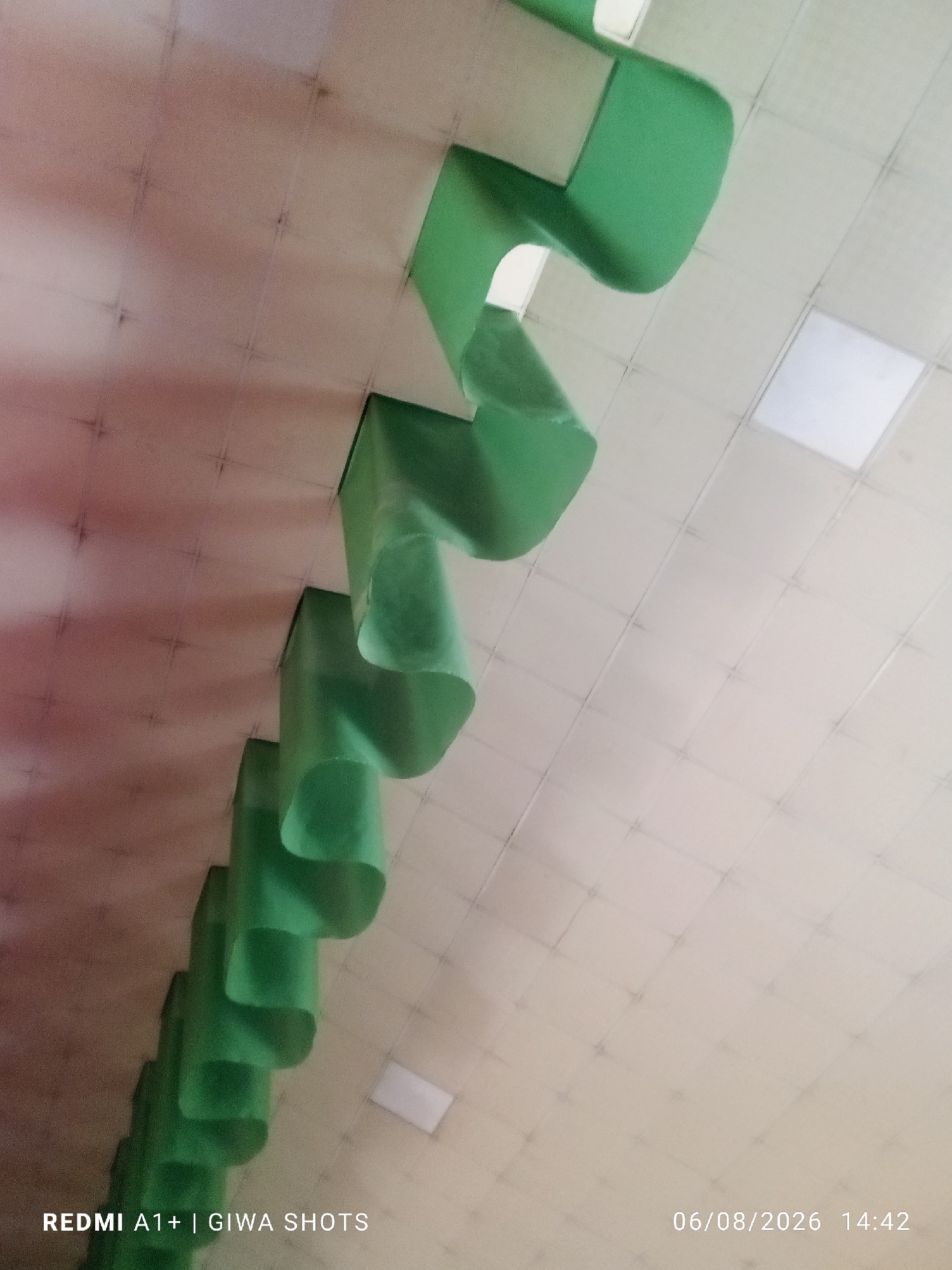 A row of green decorative elements hangs from a tiled ceiling, casting shadows. The ceiling tiles are square and arranged in a grid pattern, with visible dark lines separating them. There are some rectangular light fixtures integrated into the ceiling, one of which is brightly illuminated. The green elements appear to be made of a flexible material and are shaped into a series of curves and folds, creating a scalloped effect. The overall lighting suggests an indoor environment. The image is a low-angle shot looking upwards. The bottom of the image contains text that reads "REDMI A1+ | GIWA SHOTS" and "06/08/2026 14:42".