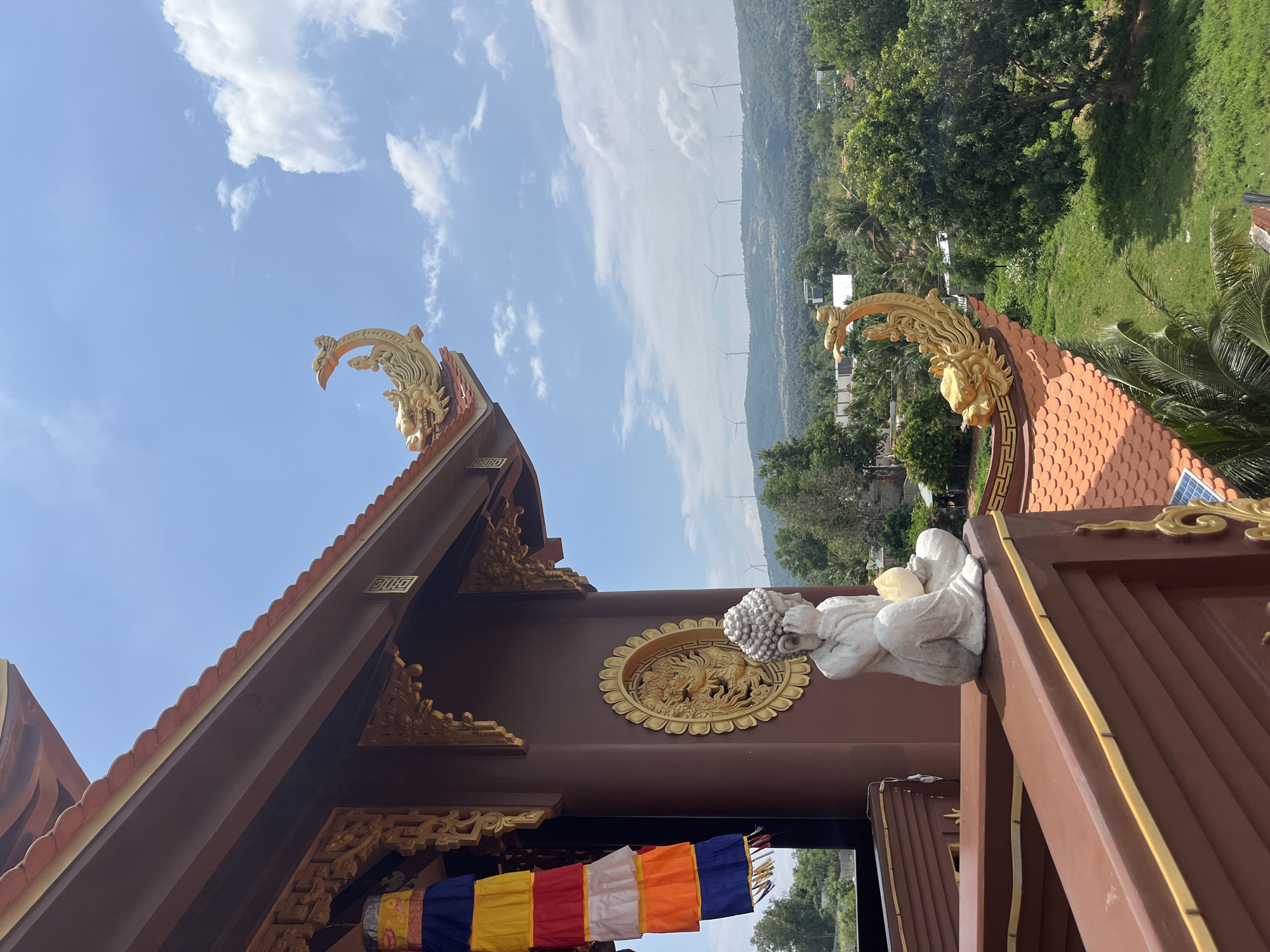 This image captures a serene view from a temple in Mũi Né, Vietnam. In the foreground, a small stone statue of Buddha sits contemplatively on a brown wooden railing, adorned with colorful prayer flags hanging to the left. The temple's architectural features are prominent, with a reddish-brown, tiled roof detailed with ornate golden dragon sculptures. Beyond the temple, a lush green landscape unfolds, dotted with trees and scattered buildings. In the distance, a line of windmills stands silhouetted against a bright blue sky with scattered white clouds, suggesting a breezy and open environment. The overall scene evokes a sense of peace and spirituality amidst a picturesque natural setting.