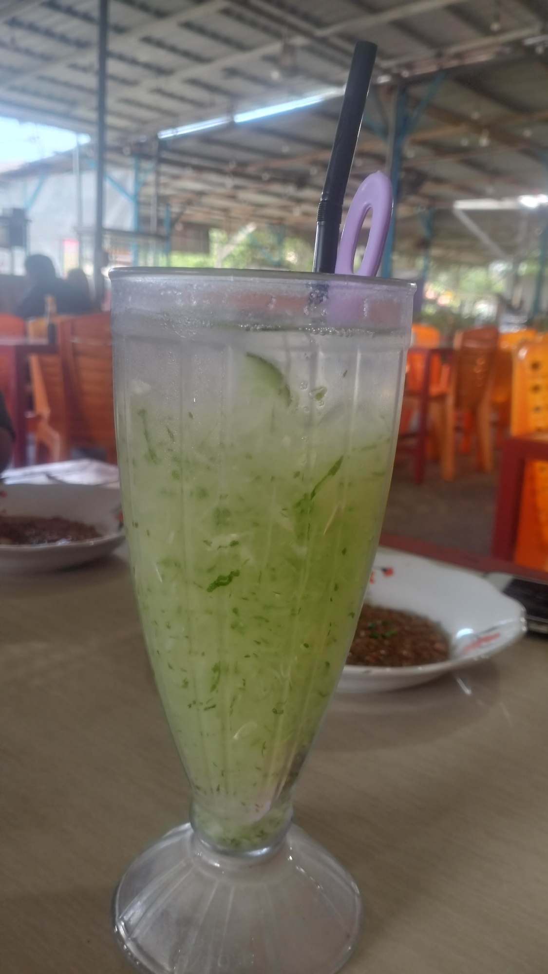 A tall, clear glass filled with a light green, icy beverage dominates the center of the frame, resting on a light-colored wooden table. The drink appears to be a refreshing concoction, likely a cucumber or melon-based drink, possibly similar to Es Timun Suri or Es Kuwut, popular in Indonesia. It contains numerous small, grated pieces of the green ingredient and ice cubes, with visible condensation on the outside of the glass, indicating its cold temperature. A black plastic straw is inserted into the drink, accompanied by a small, light purple, oval-shaped plastic clip or straw holder attached to the rim of the glass.
In the blurred background, signs of a casual dining establishment are visible. To the left and right, parts of white plates with brown, textured food (possibly savory dishes) can be seen, suggesting a meal is in progress or has just concluded. Beyond the table, the setting appears to be a covered, open-air eating area, possibly a restaurant or food stall in Lhokseumawe, Indonesia. Numerous orange and red plastic chairs and wooden tables are scattered throughout the space. The overhead structure features a metallic, corrugated roof supported by light blue and grey beams, typical of such establishments. Bright natural daylight streams in from the open sides, indicating it is daytime. A blurry figure of a person is partially visible seated in the far left background. No specific text is visible in the image. The overall impression is one of a relaxed, casual dining experience under the bright Indonesian sun.