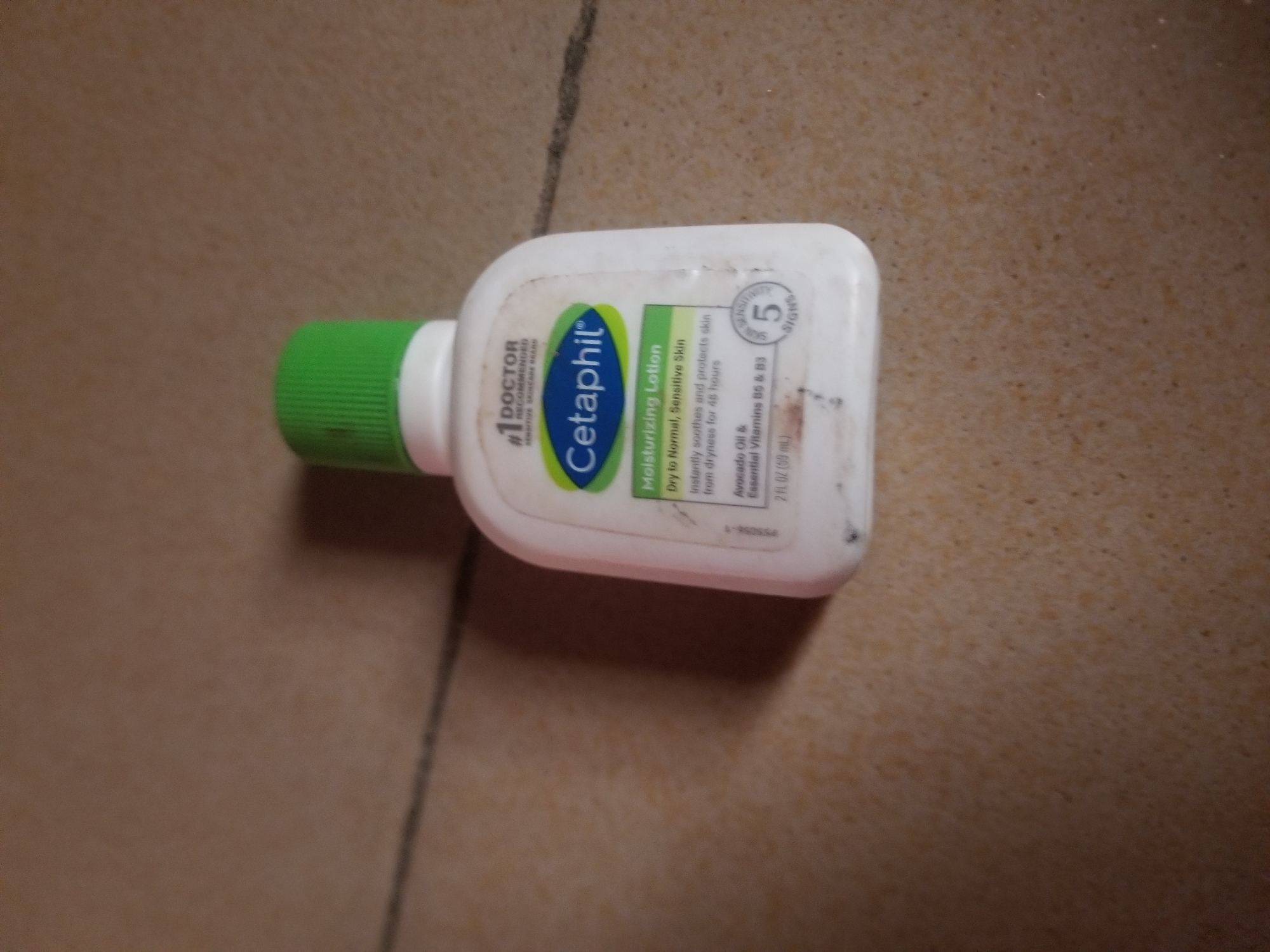 The image shows a small white bottle of Cetaphil Moisturizing Lotion with a green cap, lying on a beige tiled surface. The bottle is positioned diagonally. Visible text on the label includes "#1 DOCTOR RECOMMENDED SENSITIVE SKINCARE BRAND", "Cetaphil", "Moisturizing Lotion", "Dry to Normal, Sensitive Skin", "Instantly soothes and protects skin from dryness for 48 hours", "Avocado Oil & Essential Vitamins B5 & B3", and "2 FL OZ (59 mL)". There is also a circular graphic with the number "5" and the word "SIGNS" within it. The lighting suggests it is daytime, and the indoor setting appears to be a typical room. There are no people or specific activities depicted. The overall impression is of a product shot, possibly for sale or review, set against a simple background. The location context of Jauro Yinu, Nigeria, is not visually evident from the image itself but is provided as additional information.
