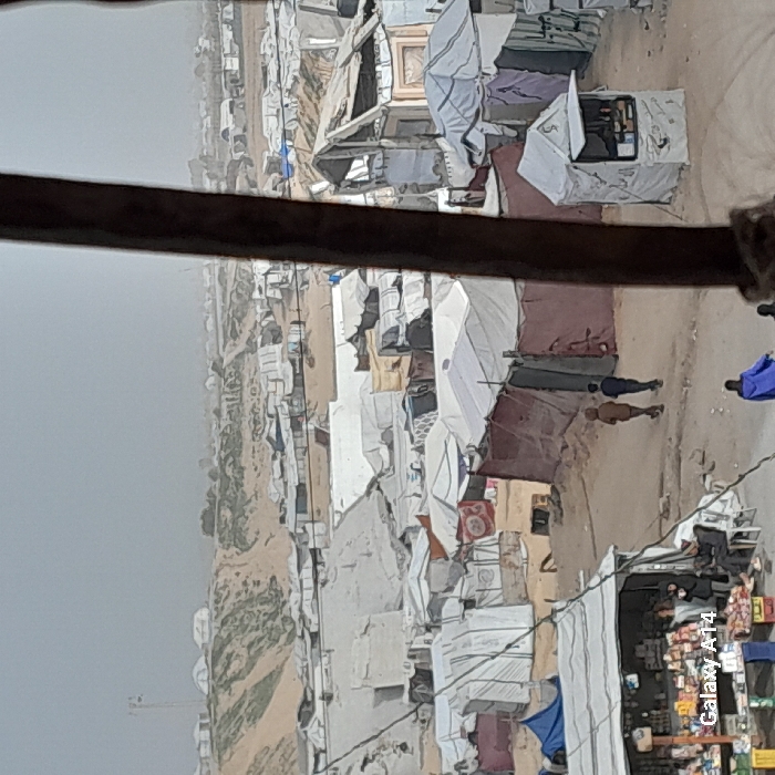 The image captures an expansive, elevated view of a densely populated, makeshift settlement or refugee camp in Khan Yunis, Palestinian Territory. The scene is dominated by numerous temporary structures, constructed from a variety of materials including white and colored tarps, fabric sheets, and corrugated metal, indicating challenging living conditions.
A dark, horizontal bar partially obstructs the foreground, suggesting the photo was taken from an elevated vantage point, possibly from within a building or through a window. Below this bar, a network of narrow, unpaved paths or alleys winds through the camp.
Several people are visible within the camp. In the middle ground, two individuals are walking along a dirt path. One wears light-colored clothing, while the other is in darker attire. Further to the right, another person is seen wearing a bright blue garment. On the far right side of the image, a small, makeshift market stall or shop displays various goods on shelves and tables, suggesting economic activity within the camp. Clothes are also visible hanging to dry on several of the shelters.
The background reveals more layers of similar temporary dwellings stretching into the hazy distance, underscoring the vastness of the settlement. The ground appears dry and dusty, with minimal vegetation. The sky is overcast or hazy, diffusing the daylight and preventing strong shadows, which suggests either an overcast day or significant dust in the air. The overall atmosphere conveys a sense of hardship and crowded, temporary living.
A visible watermark, "Galaxy A14," is present in the bottom right corner of the image.