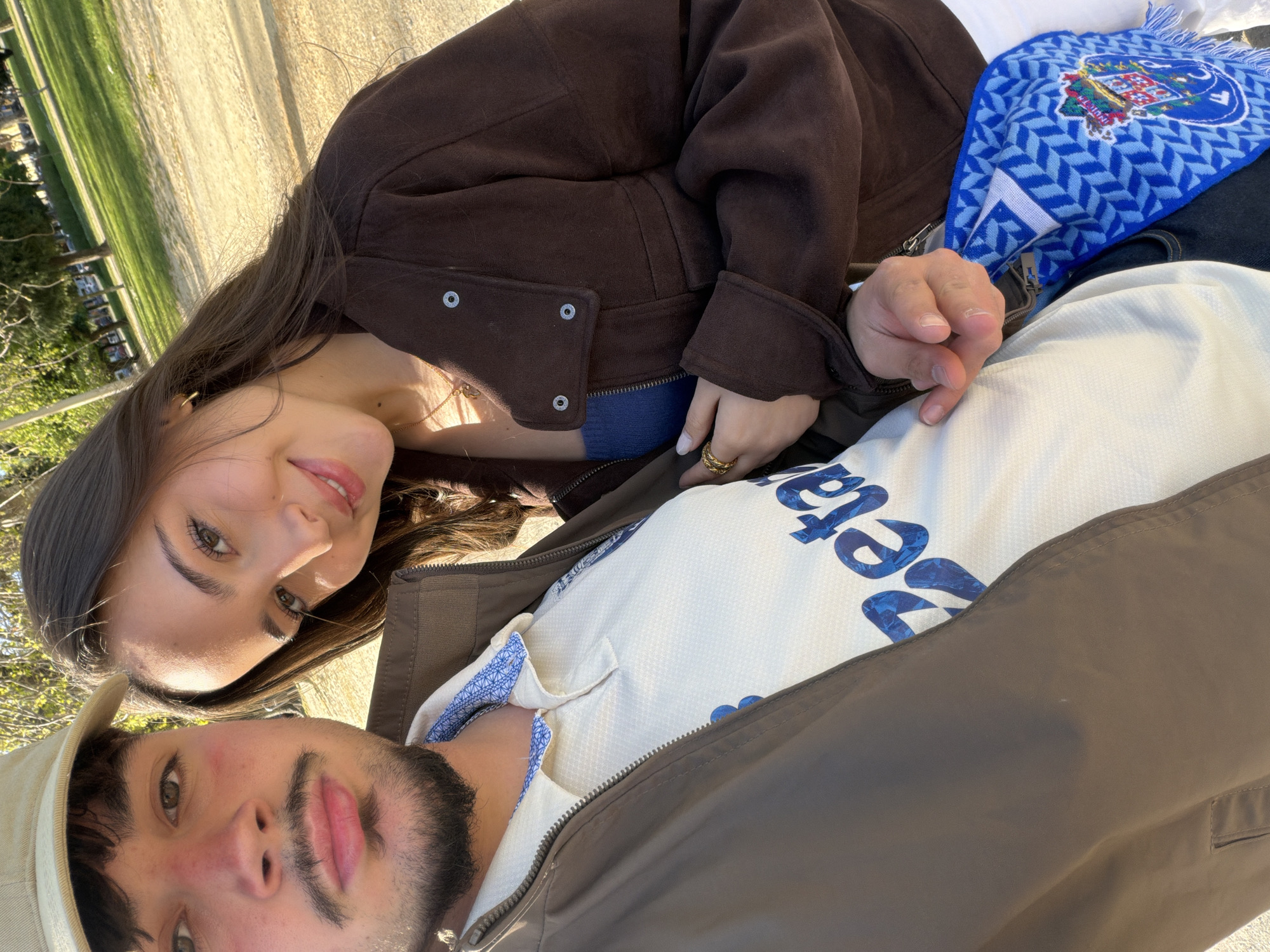 A couple is taking a selfie on a sunny day, possibly in Porto, Portugal, based on the Porto soccer scarf draped over the man in the foreground. The woman is on top, wearing a brown jacket and a gold ring, her face visible and turned towards the camera with a slight smile. The man is wearing a beige hat and a jacket, with a shirt underneath that has blue lettering. The background shows a park-like setting with green grass and trees, with a sandy or concrete path visible in the distance.